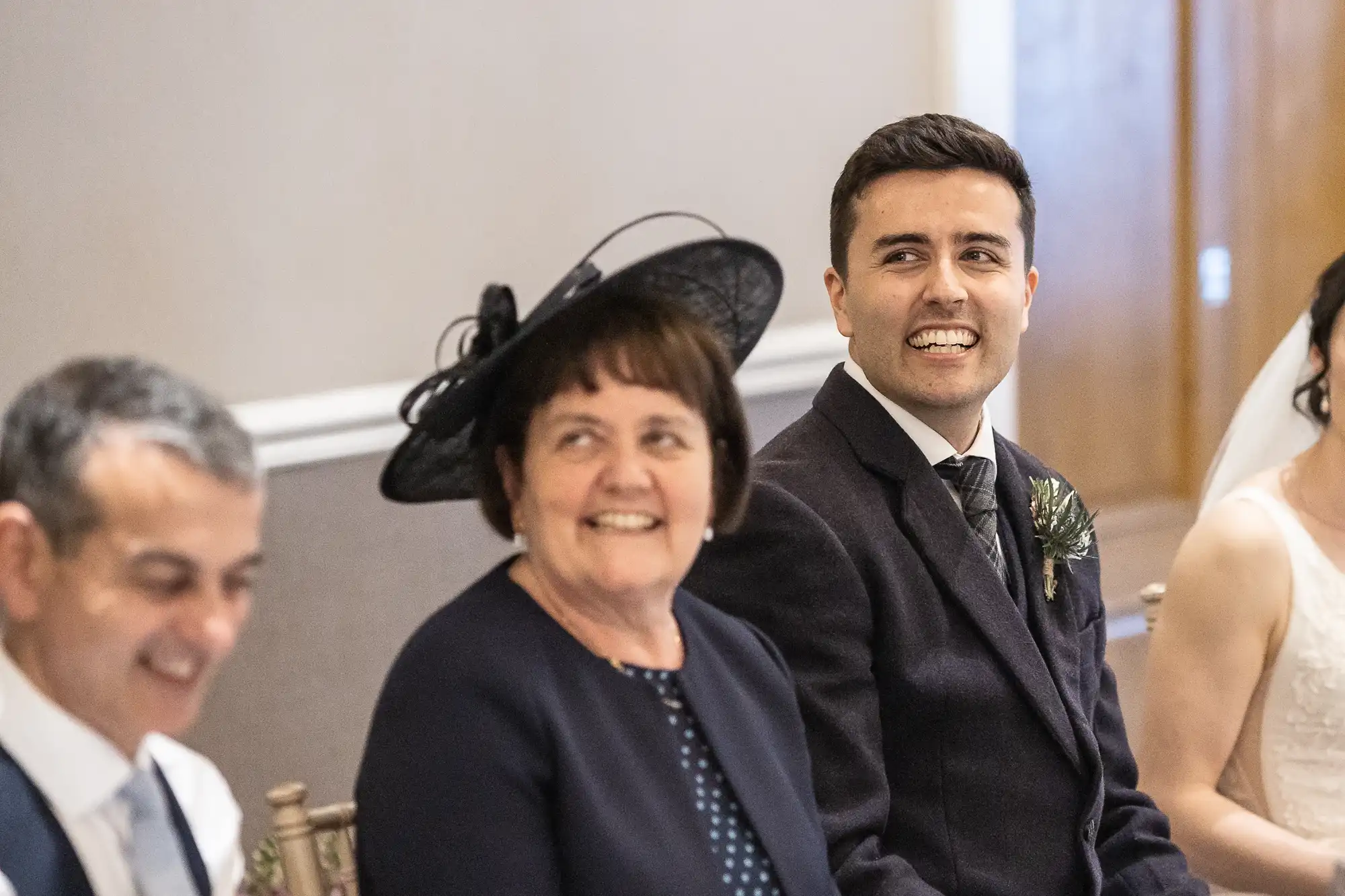 A group of people seated indoors, dressed formally. The man in the center wears a dark suit, smiling. The woman next to him is wearing a dark outfit with a decorative black hat.