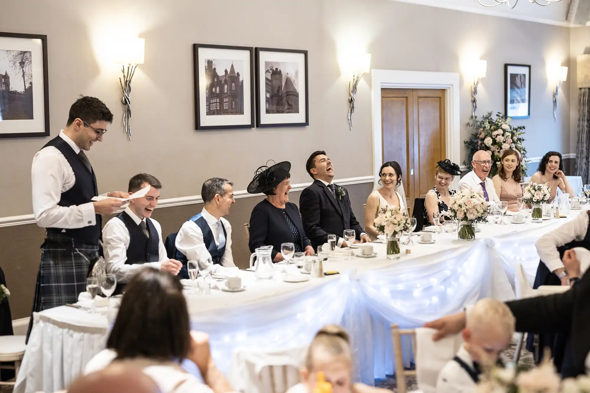 People seated at a decorated wedding reception table, listening to a standing man give a speech. One person is laughing, and floral arrangements are visible on the table.