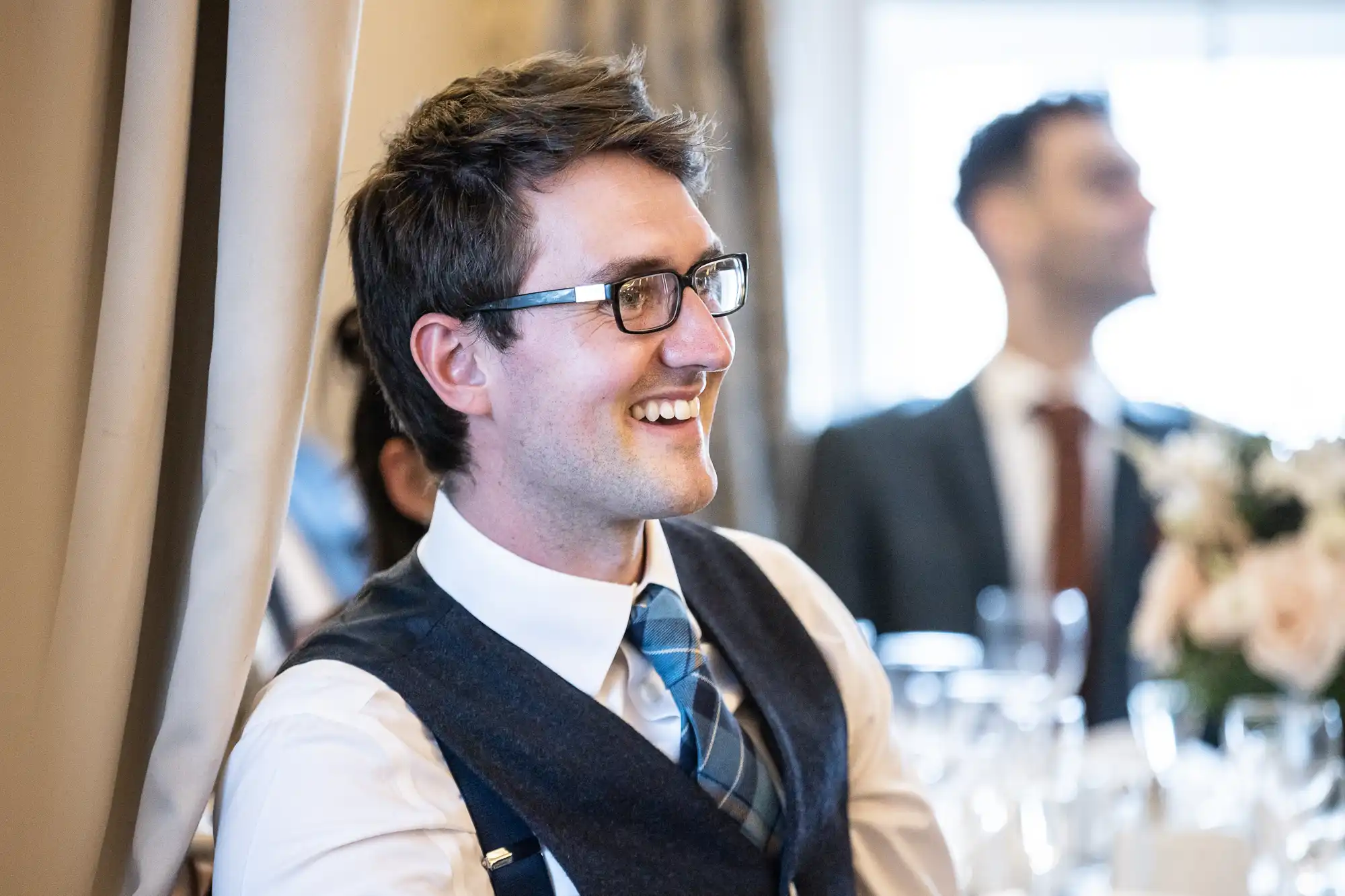 A man wearing glasses, a white shirt, and a tie smiles at an indoor event. Another person in a suit is blurred in the background.