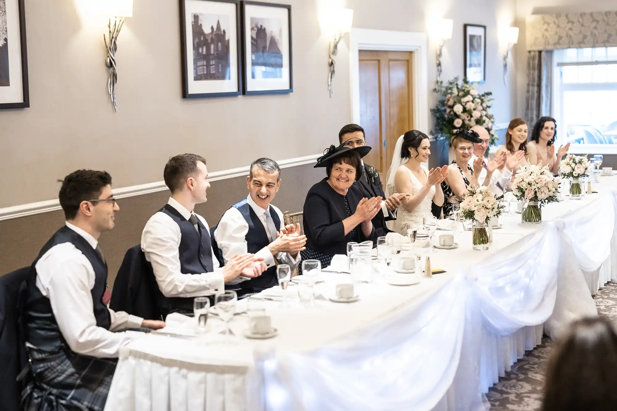 A group of people sit at a long, decorated table, clapping and smiling in a formal setting.