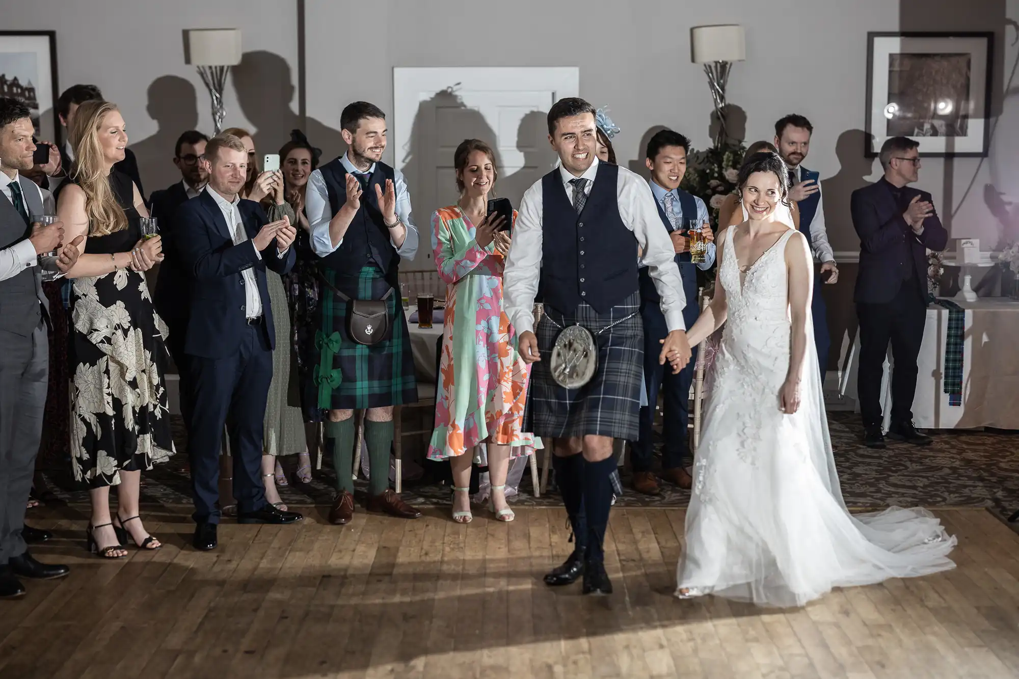 A bride and groom hold hands while walking through a crowd of applauding guests. The groom wears a kilt, and the bride is in a white dress. Background features decorations and a wooden floor.