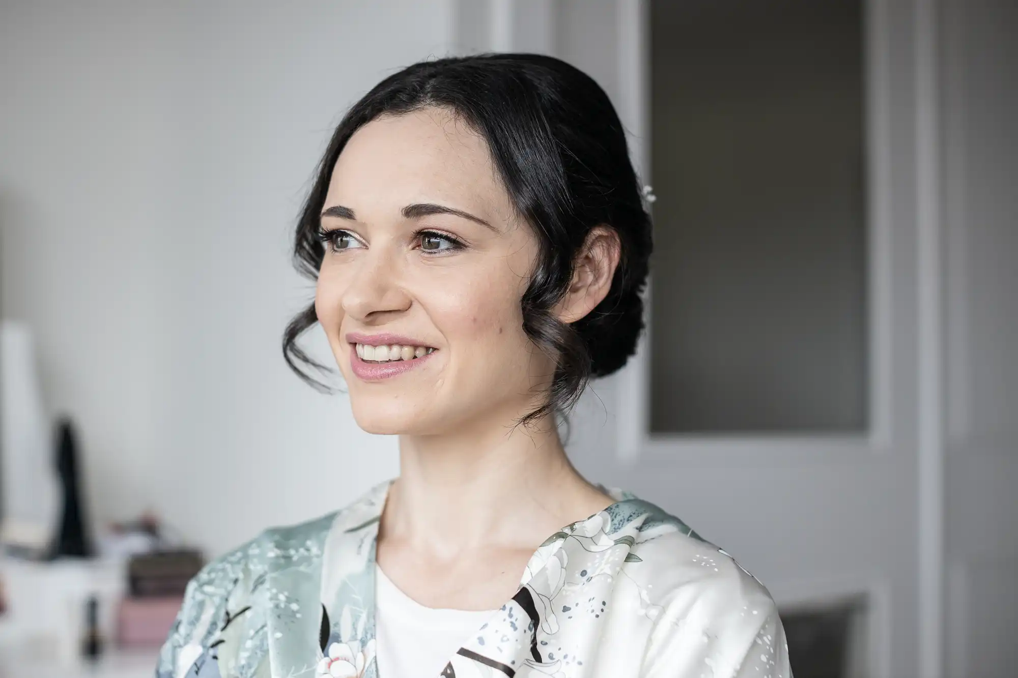 A person with dark hair smiles, looking to the side, wearing a floral-patterned outfit indoors.