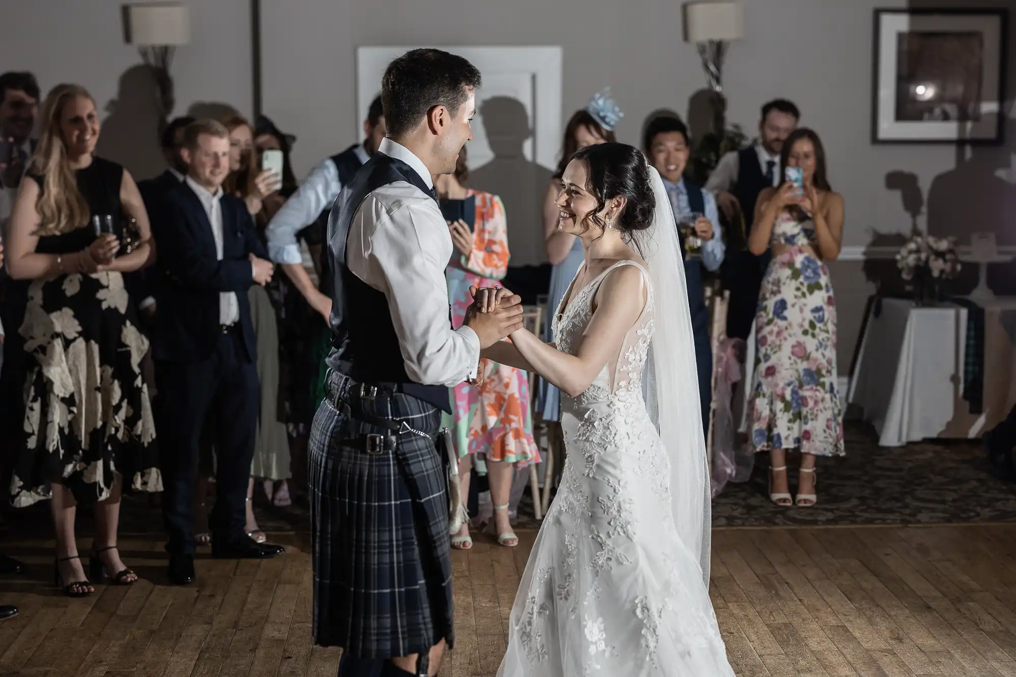Bride and groom dance in front of guests; the groom is wearing a kilt, and the bride is in a white dress with a veil. Guests watch and take photos.