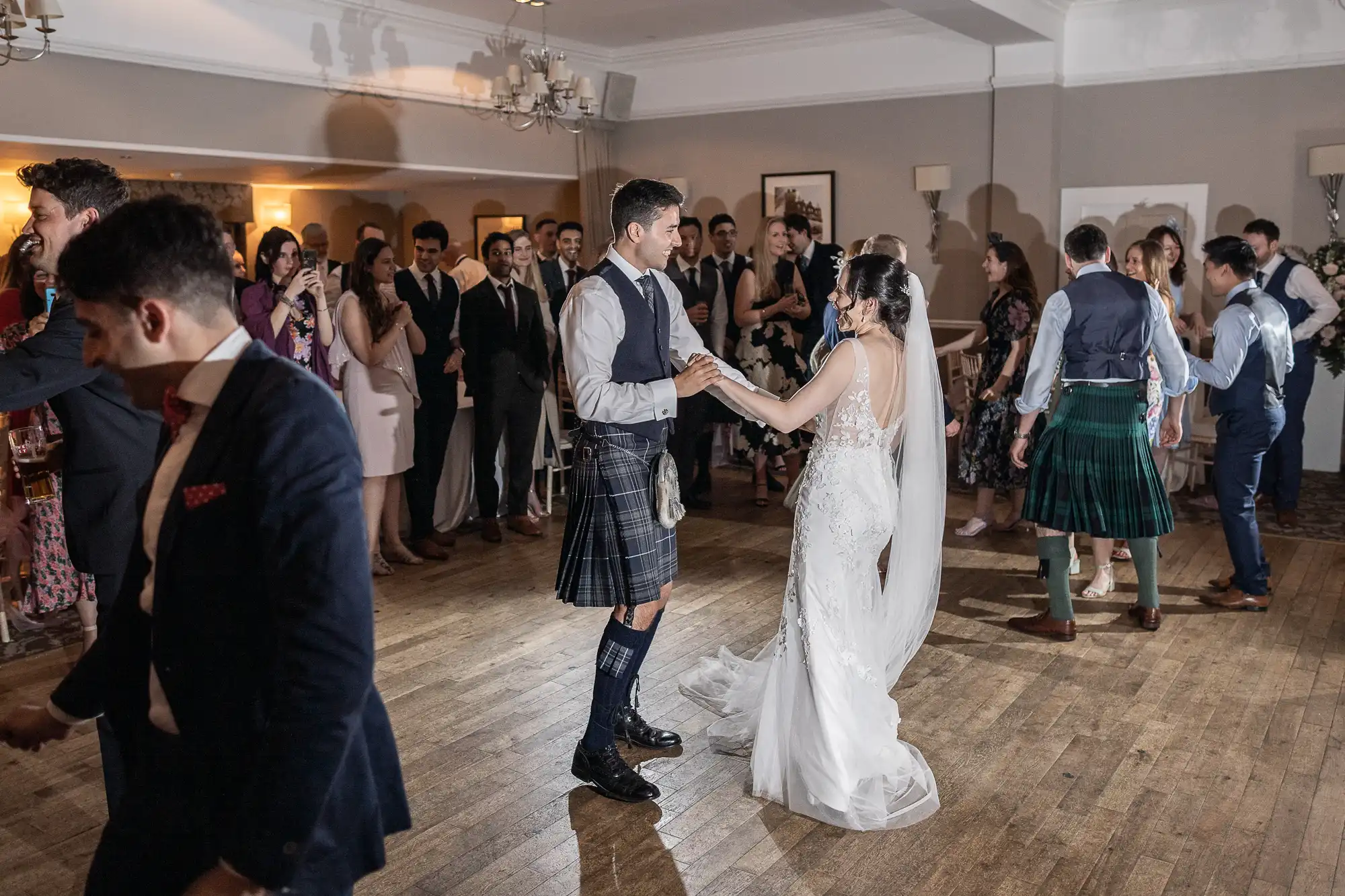 A bride and groom dance at their wedding reception, surrounded by guests. The groom wears a kilt, and the bride is in a white gown. Other guests are dancing and socializing in the background.