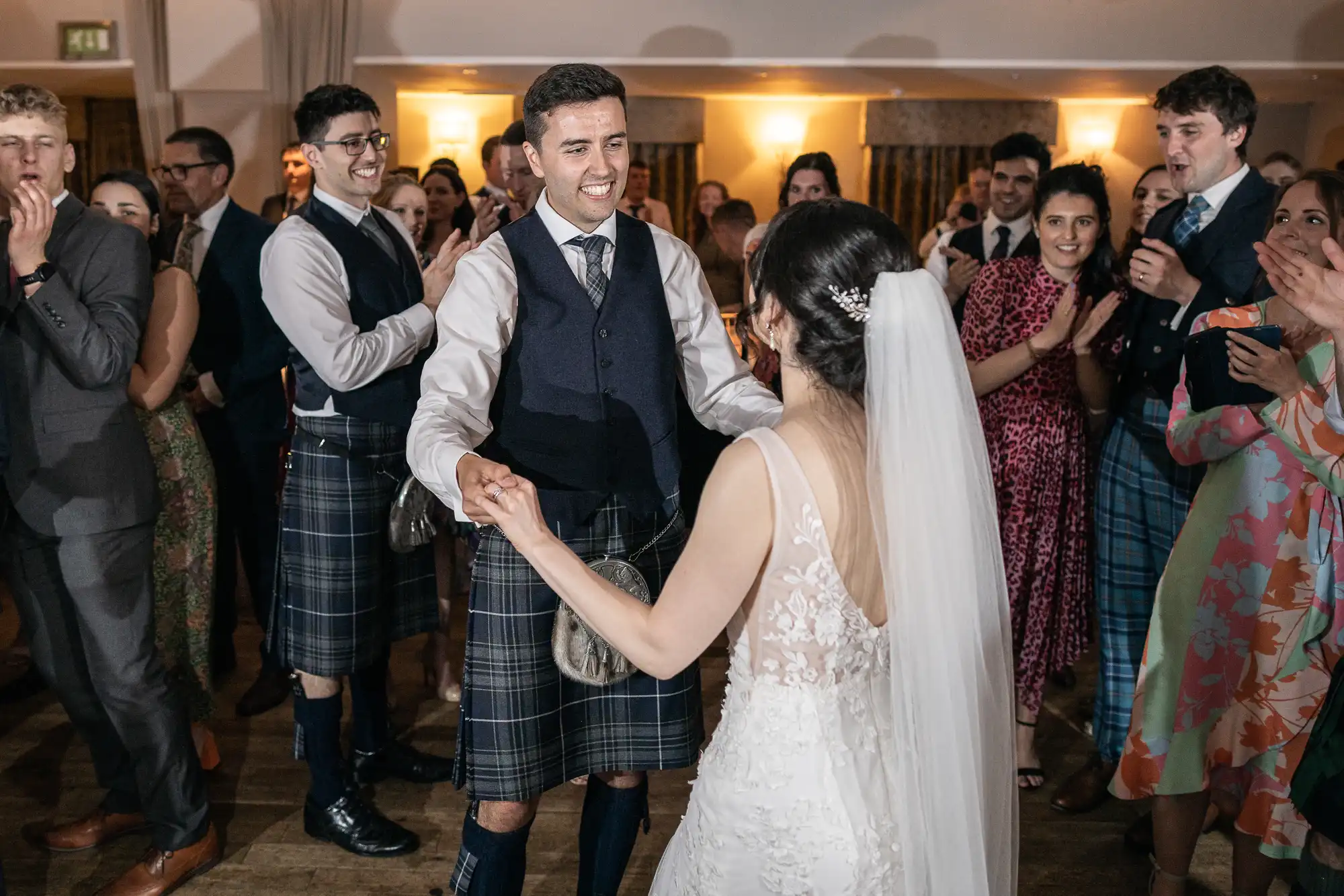 Bride and groom, surrounded by guests, dance at a wedding. Groom wears a kilt and vest, while the bride wears a white dress with a veil.