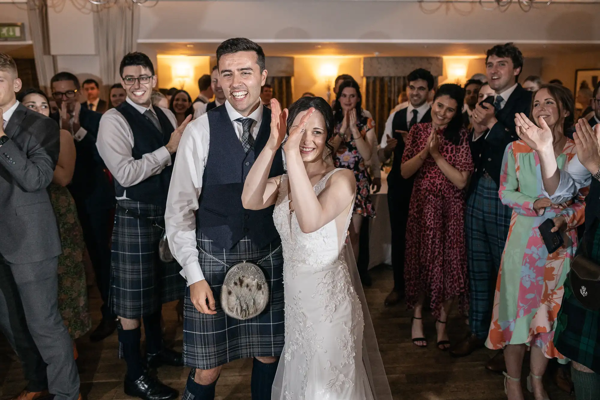 A smiling couple in wedding attire, with the groom in a kilt and the bride in a white dress, stand together while a group of guests claps and smiles around them in a warmly lit room.