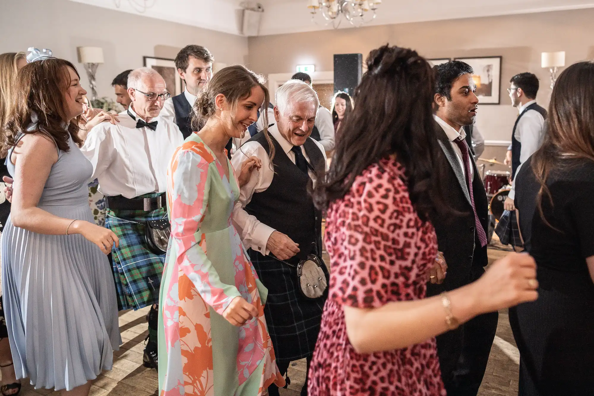 A group of people in colorful attire, some wearing kilts, are dancing together in a well-lit room.