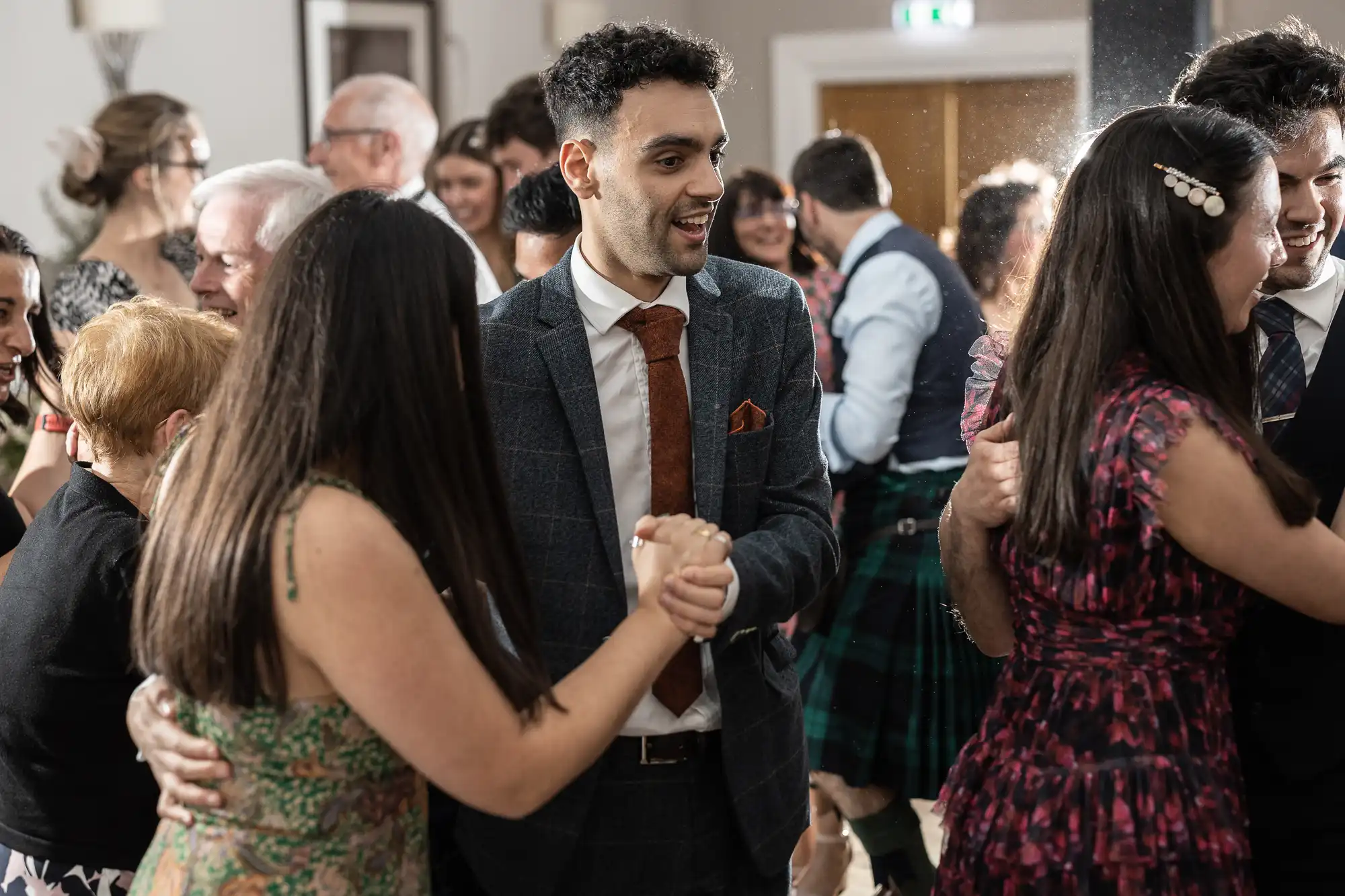A group of people dressed in formal attire are dancing and socializing at a gathering. One man in a suit is prominently visible in the center, holding hands with others.