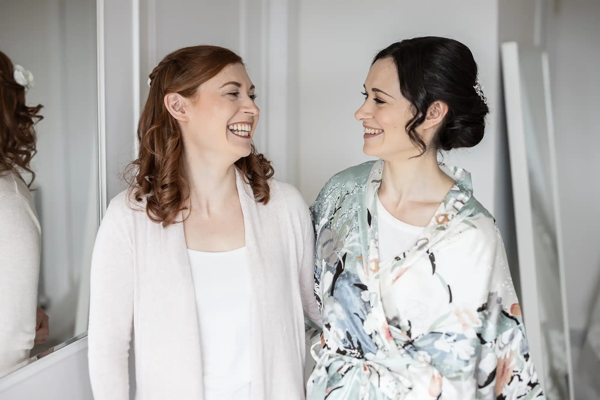 Two women smiling at each other, one in a floral robe and the other in a light cardigan, standing indoors.