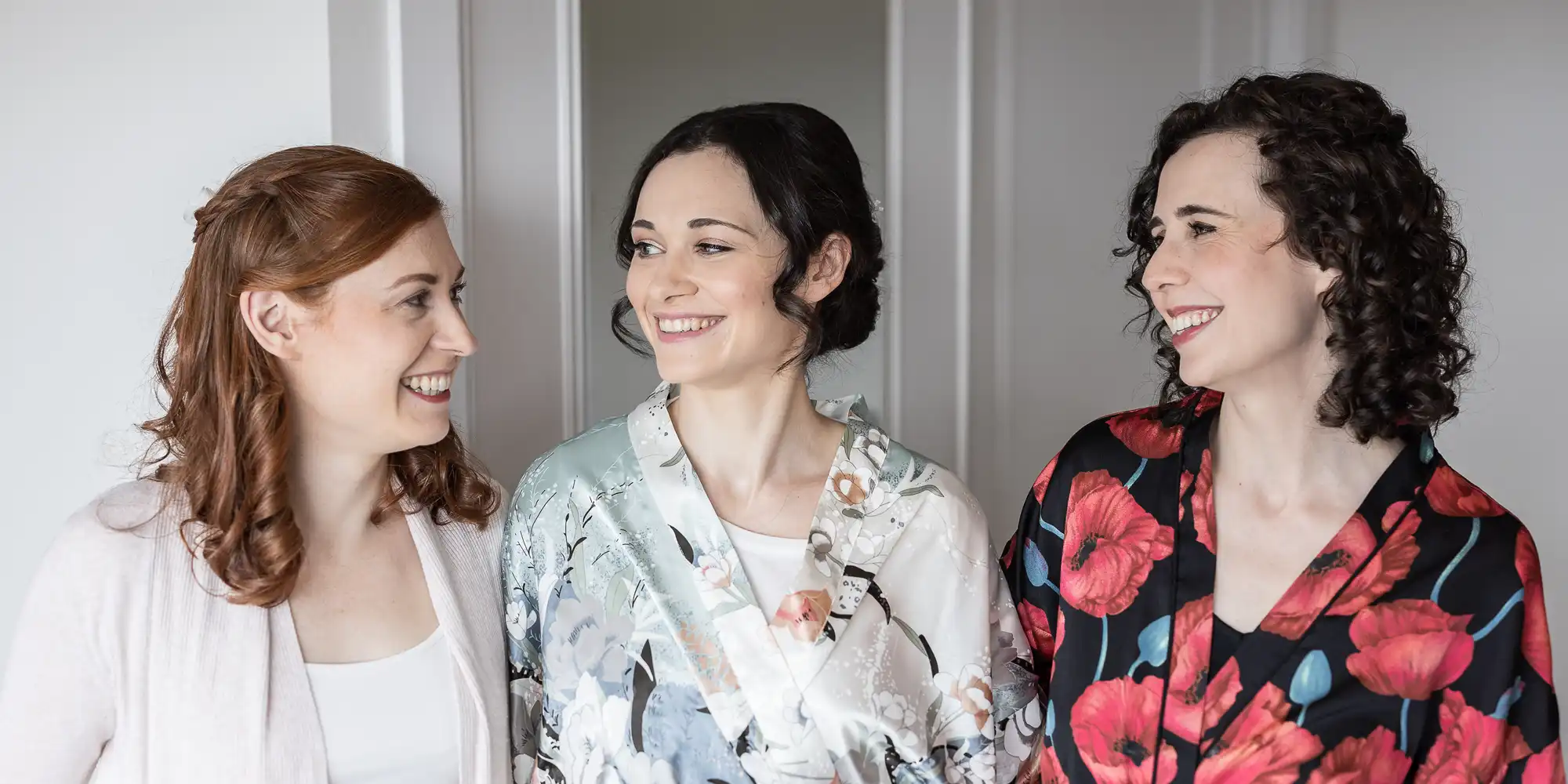 Three women smiling at each other, standing indoors, wearing floral and plain outfits.