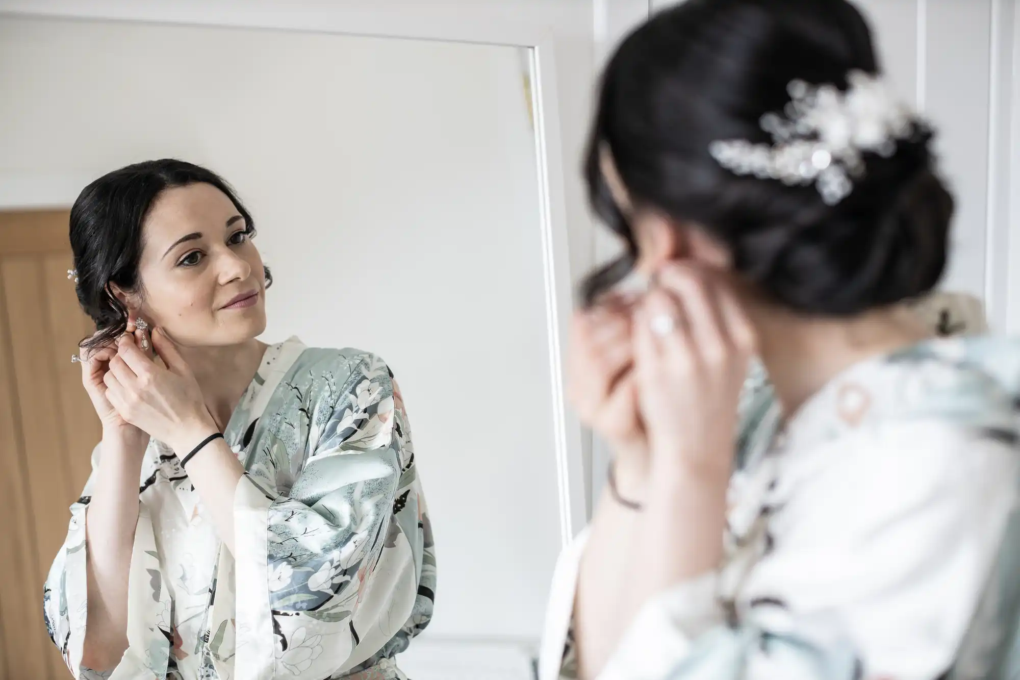 A woman in a floral robe adjusts her earring while looking in the mirror.