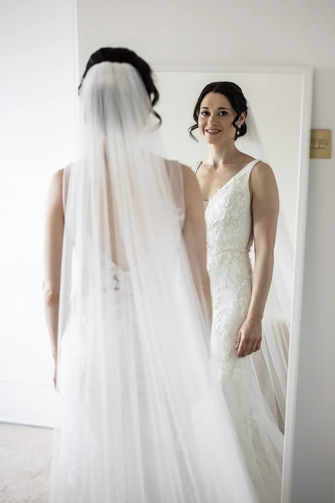 A bride in a white wedding dress and veil looks at her reflection in a mirror, smiling.