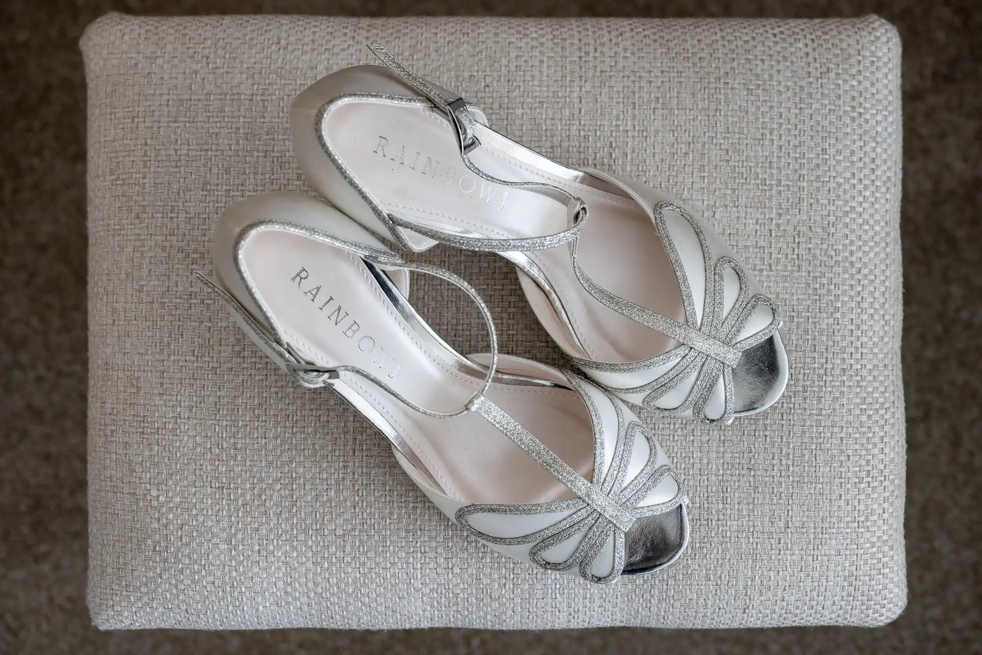 Silver peep-toe sandals with ankle straps, labeled "RAINBOW," placed on a textured beige surface.