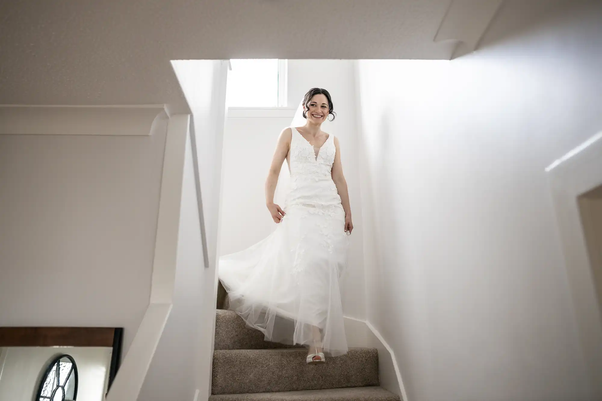 A person in a white wedding dress descends a carpeted staircase, smiling, under a bright overhead light.
