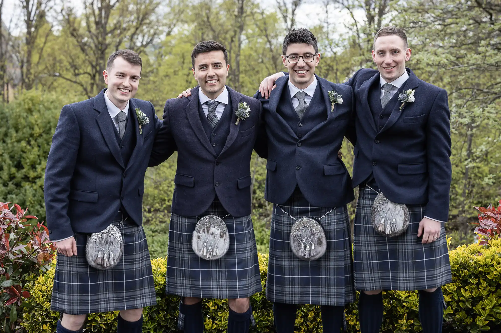 Four men in matching formal Scottish attire with kilts standing outdoors, smiling with arms around each other.