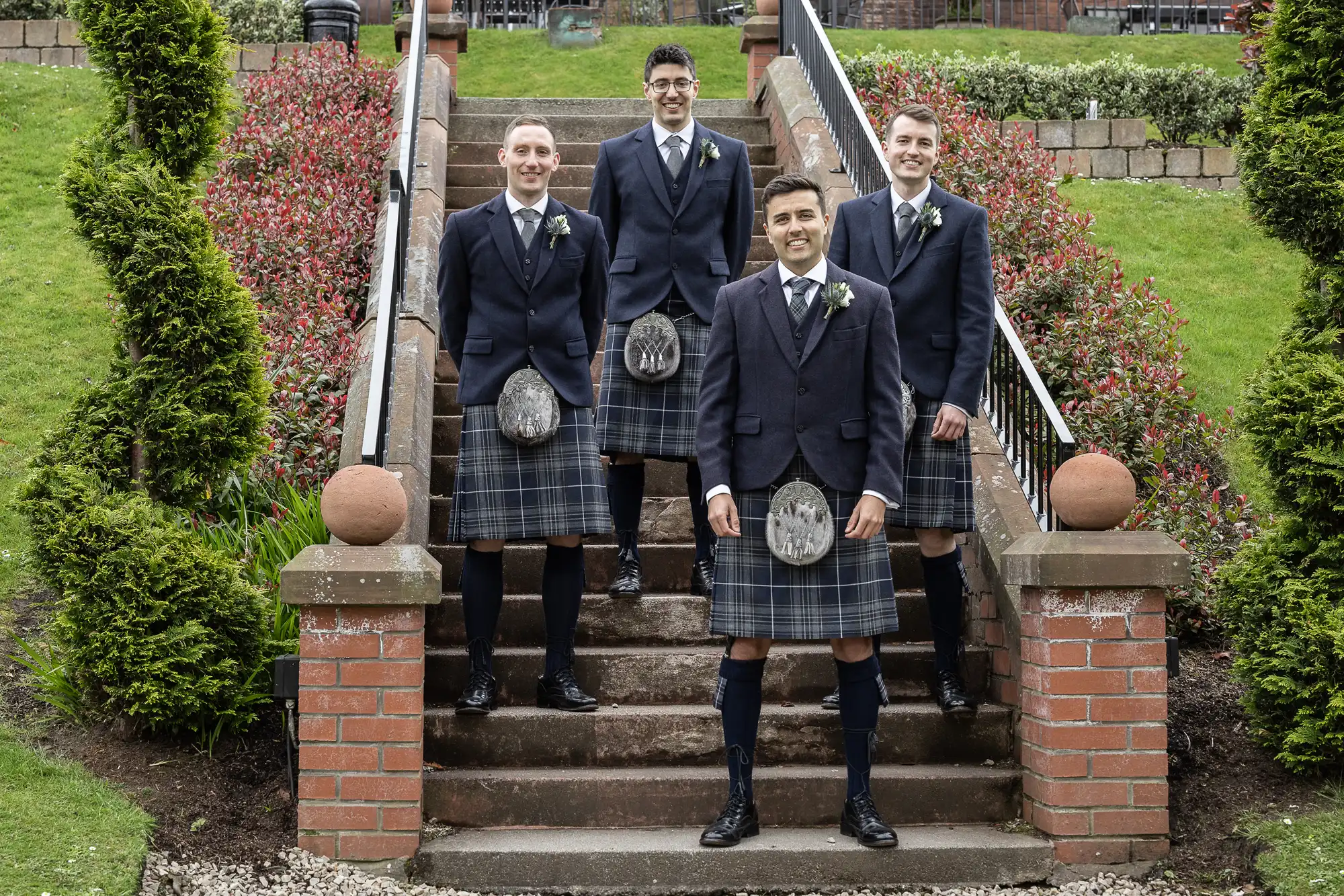 Four men in kilts and matching formal attire stand on outdoor steps, surrounded by greenery and red shrubs.