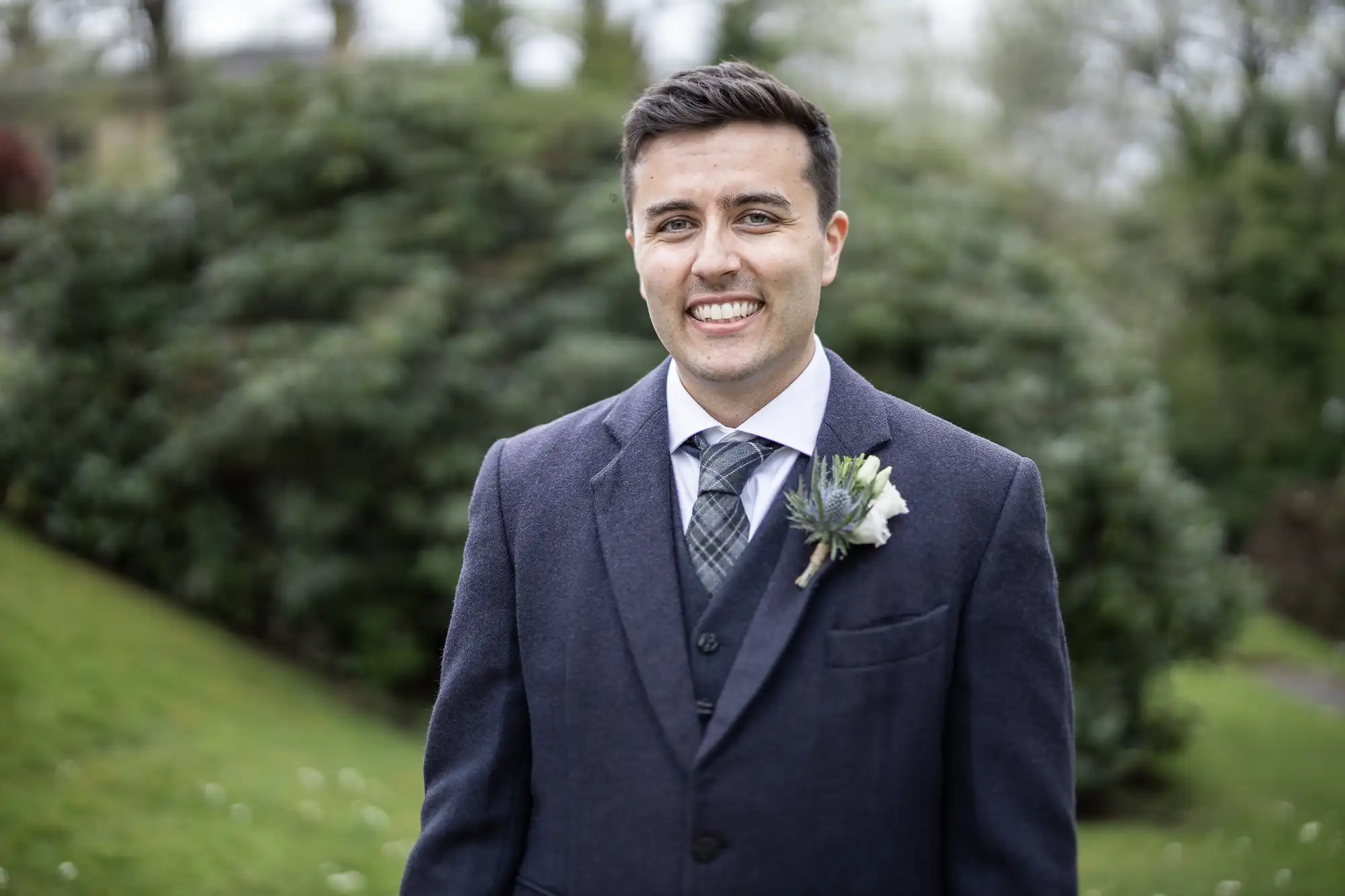 Man in a suit with a boutonniere stands outside with trees and grass in the background, smiling.