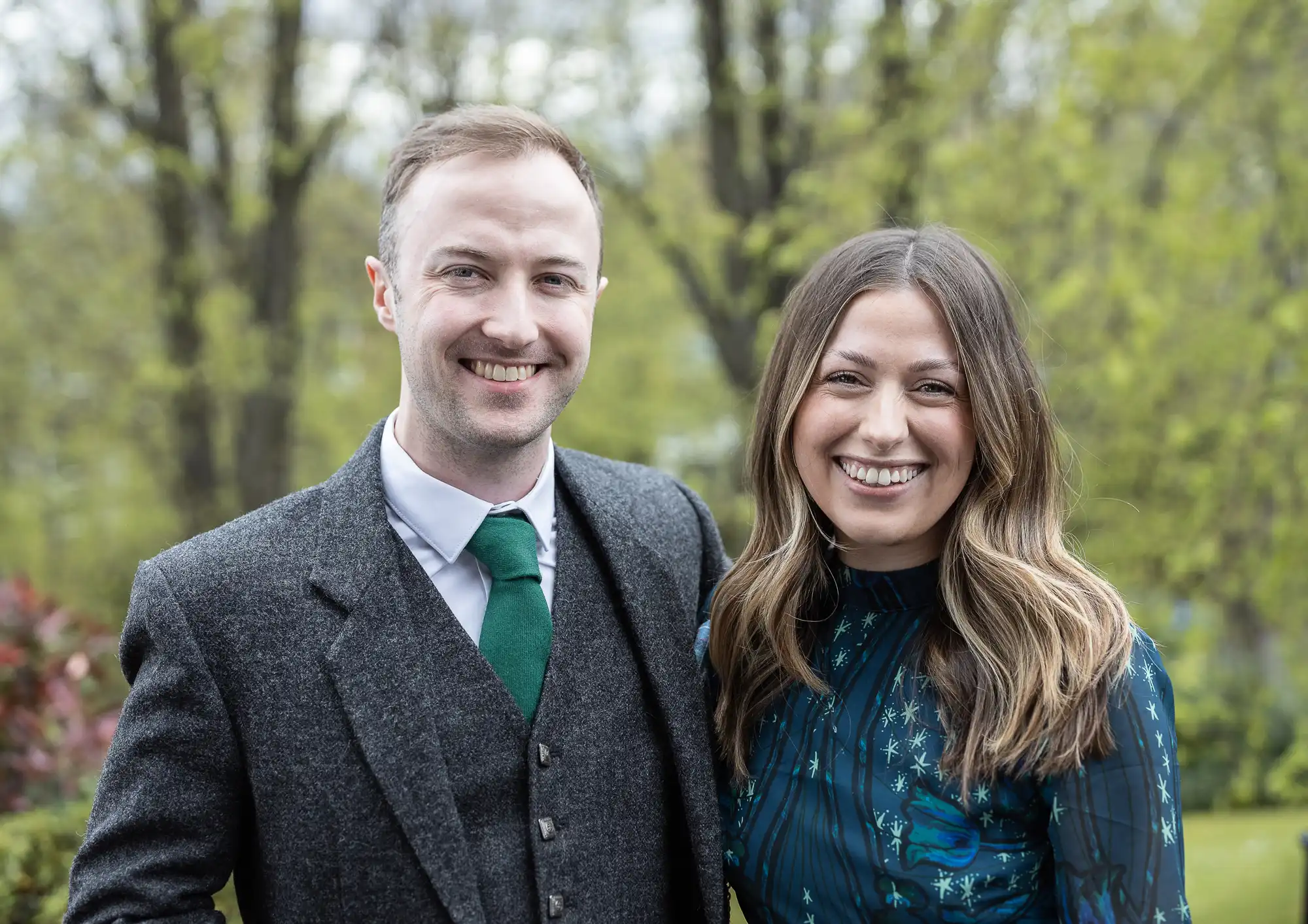 A man in a suit and a woman in a patterned dress smile while standing outside with trees in the background.