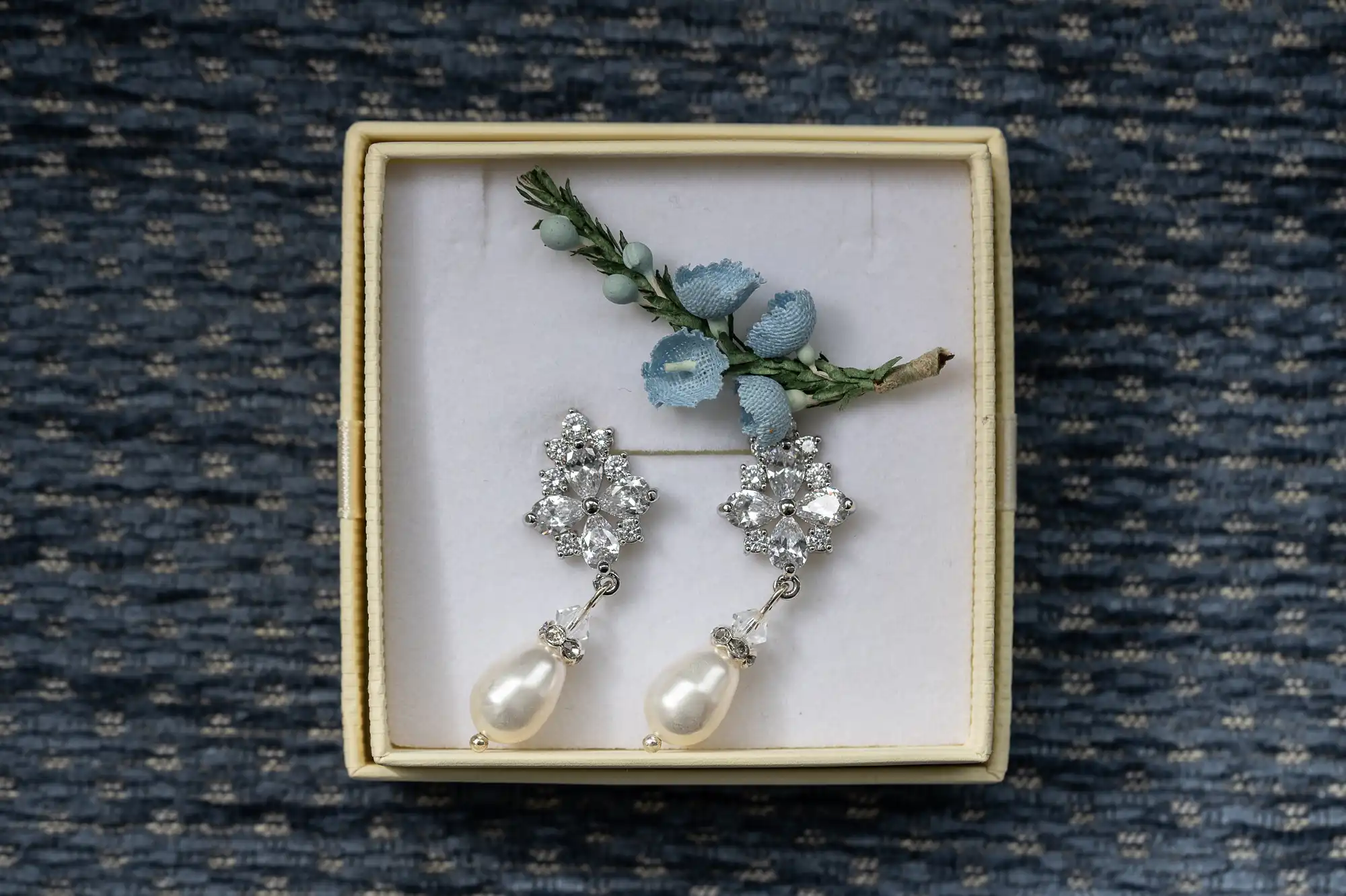 Earrings with pearl drops in a box, decorated with blue fabric flowers and green leaves on a woven fabric background.