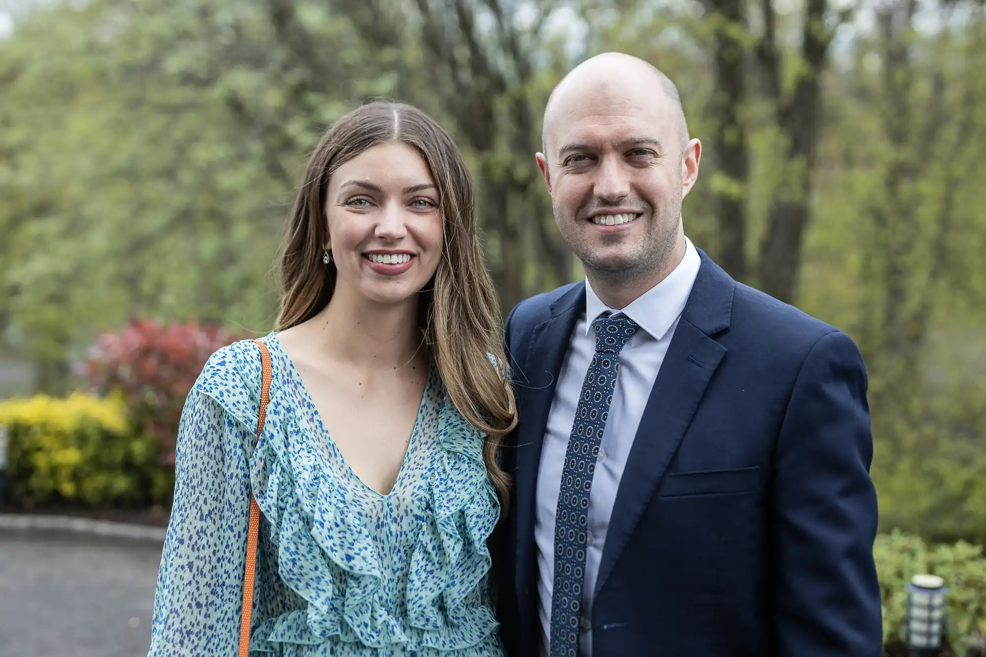 A woman in a patterned dress stands next to a man in a suit and tie, smiling outdoors with trees in the background.