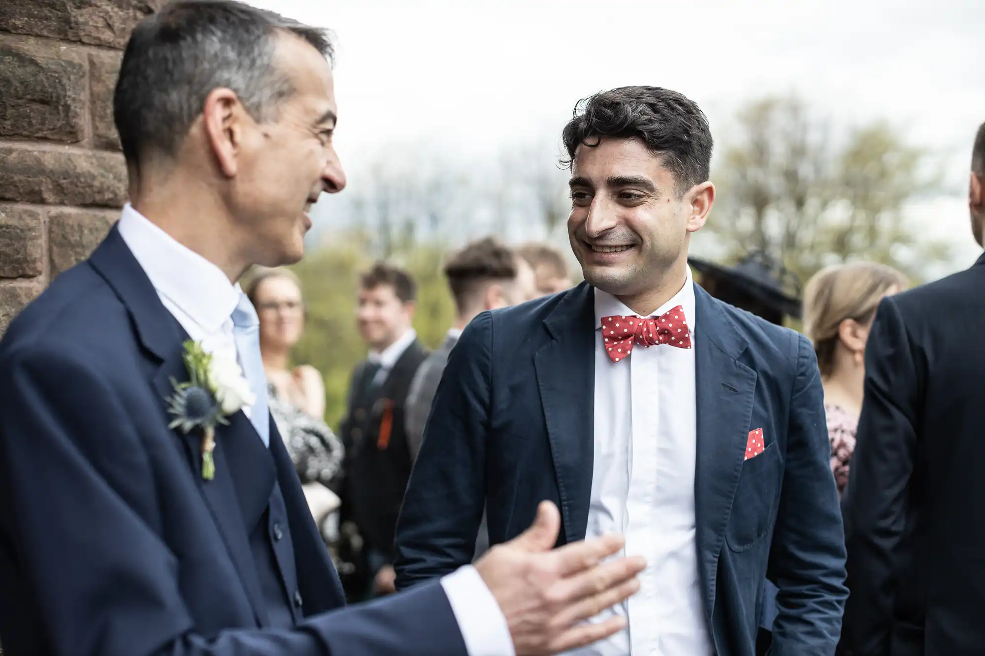 Two men in suits, one with a bow tie, are talking outside at an event. Other people are in the background.