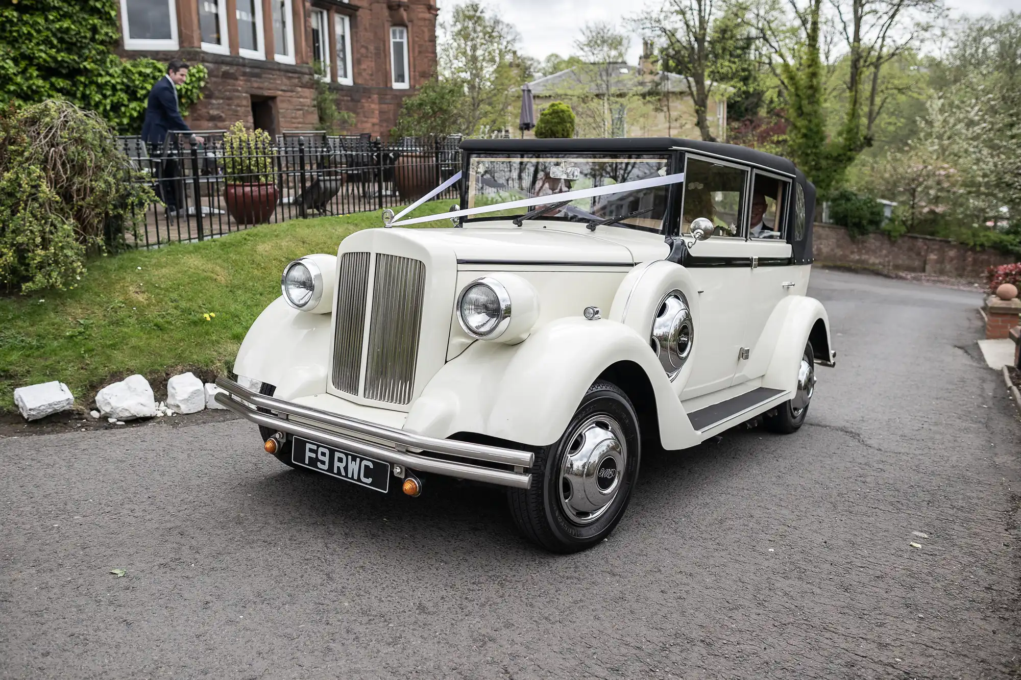 A vintage white wedding car with a ribbon on the front is parked on a driveway, surrounded by greenery.