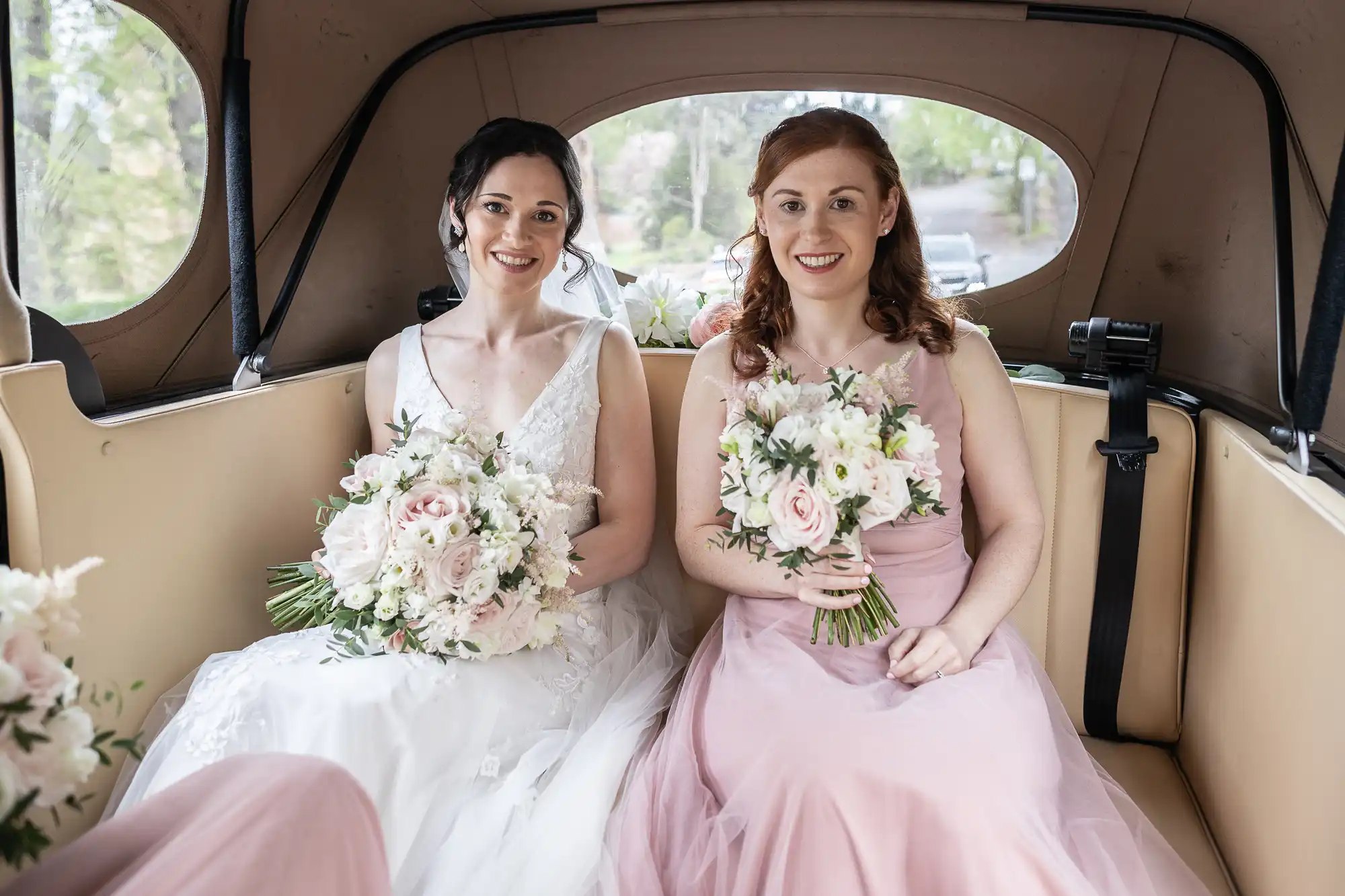 Two women in formal dresses, holding bouquets, sit in the back of a vehicle with a neutral interior.