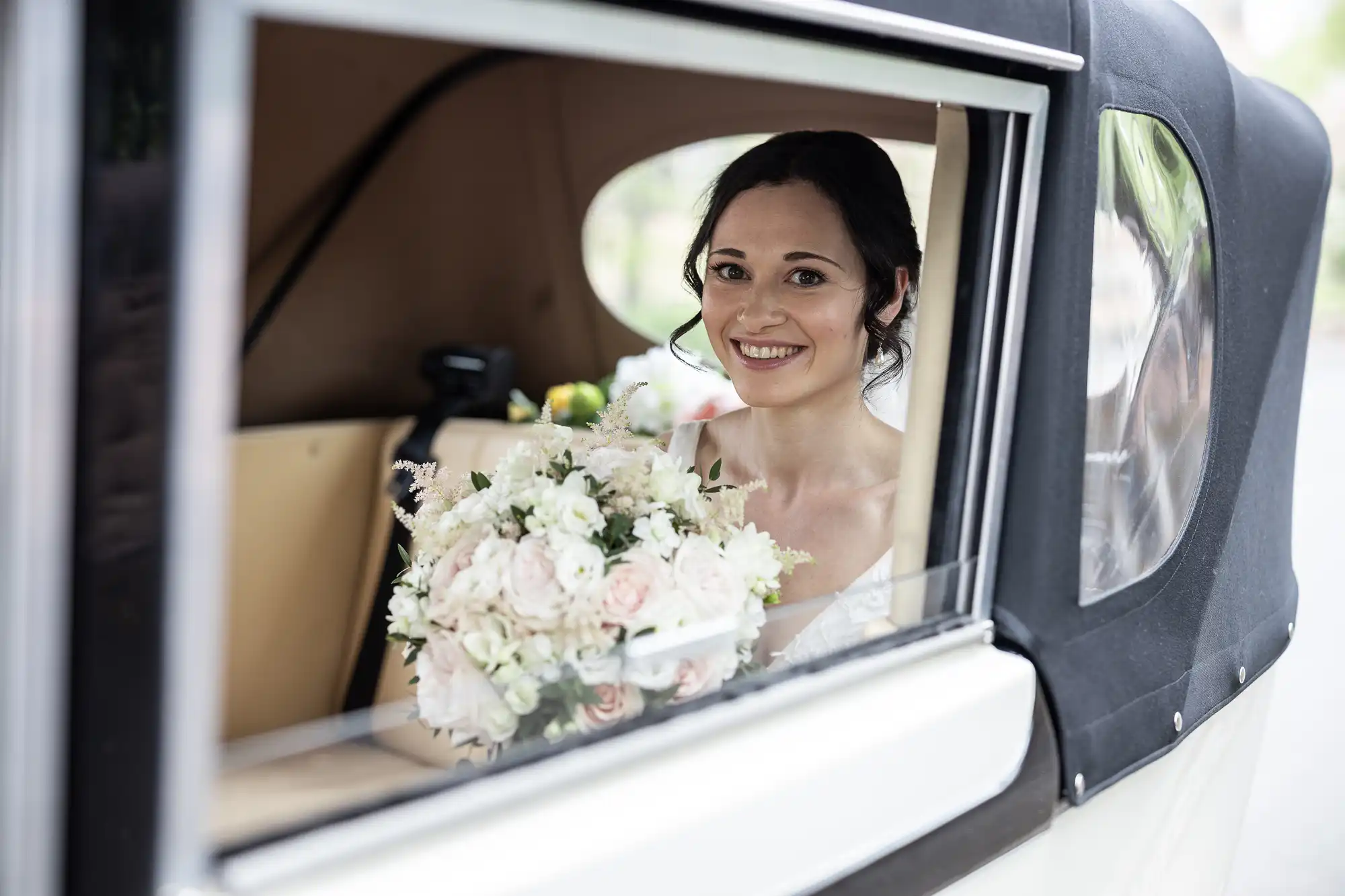 Bride smiling while sitting inside a car, holding a bouquet of white and pink flowers, with a beige interior visible through the open window.