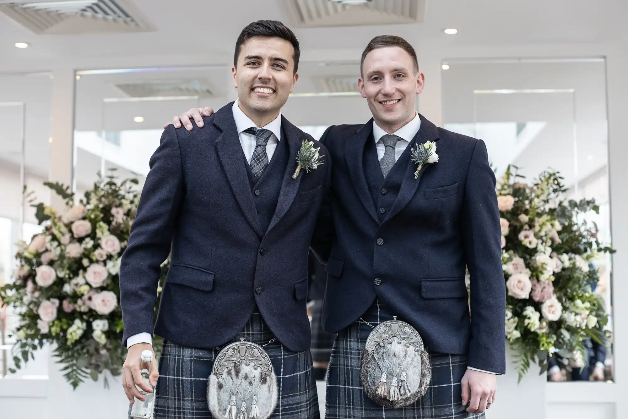 Two men in dark suits with plaid kilts, smiling and standing together indoors, with large floral arrangements in the background.