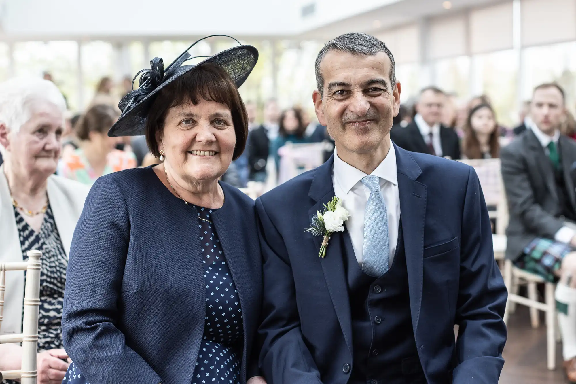 A woman in a blue outfit and hat sits next to a man in a suit with a boutonniere. They are seated at an indoor event with people in the background.