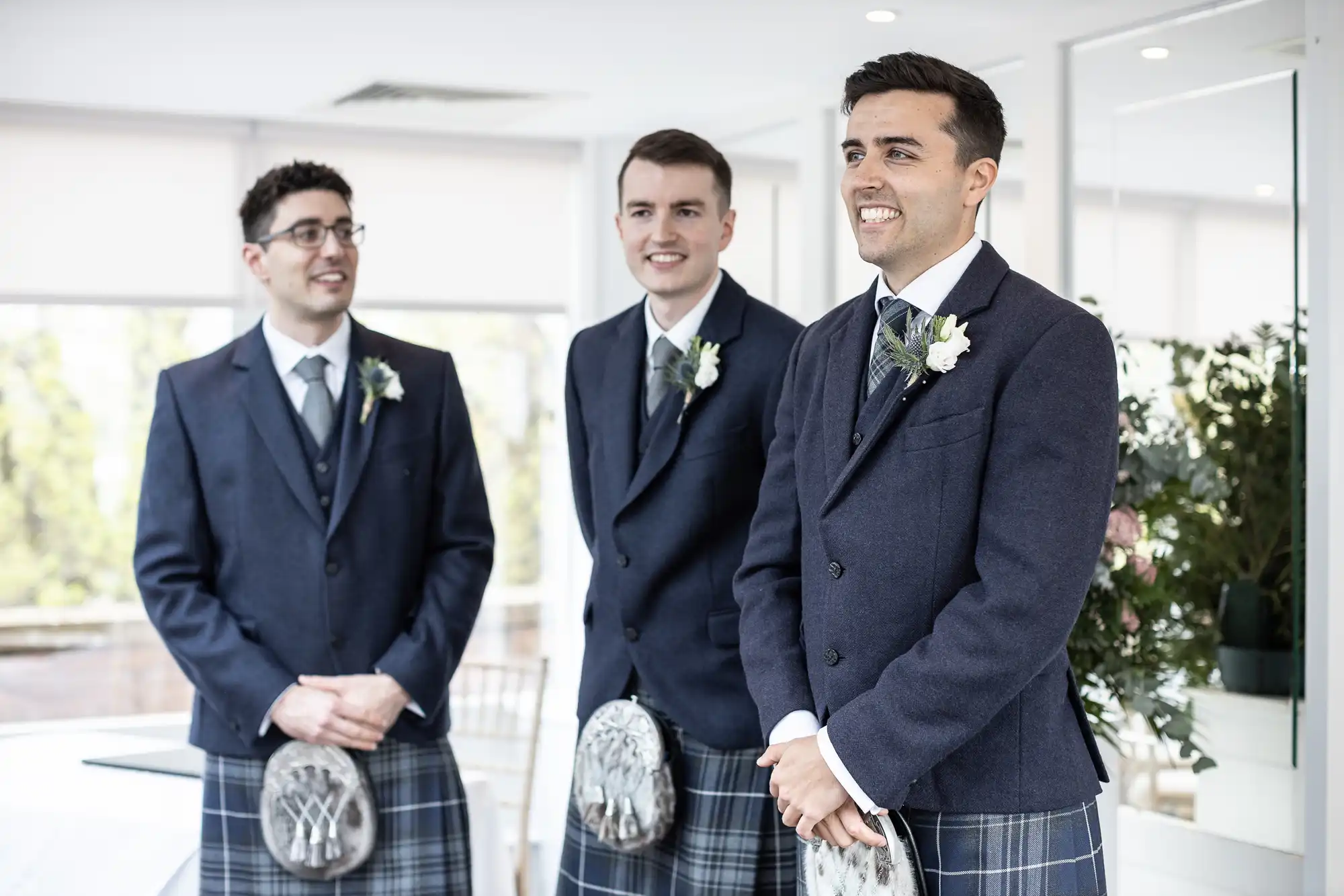 Three men in matching blue suits and kilts, each with a sporran, stand indoors smiling. They wear white shirts and ties, while two have glasses. A plant and bright windows are in the background.