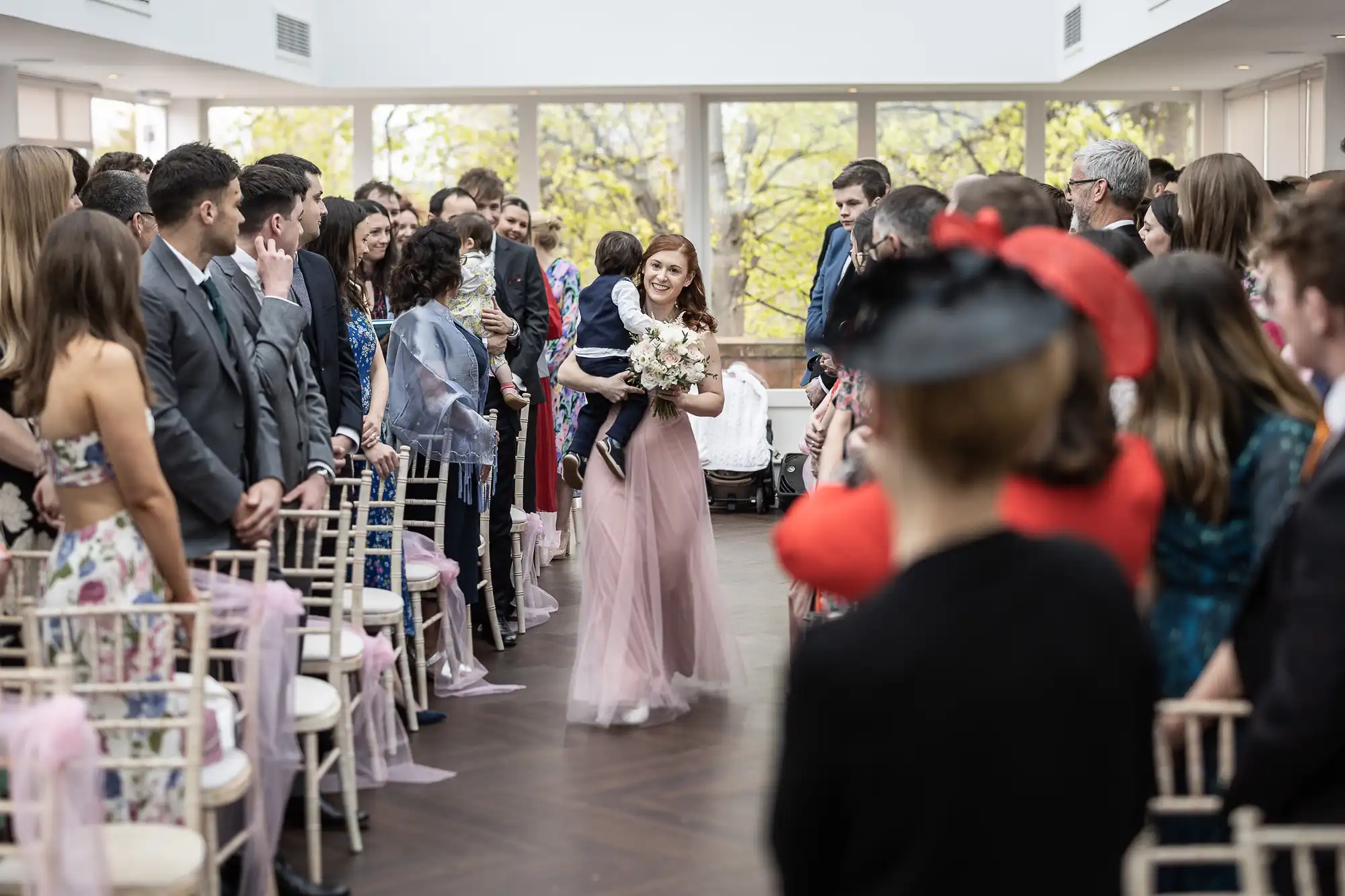 A woman in a pink dress walks down the aisle holding a bouquet, surrounded by guests in a bright room.