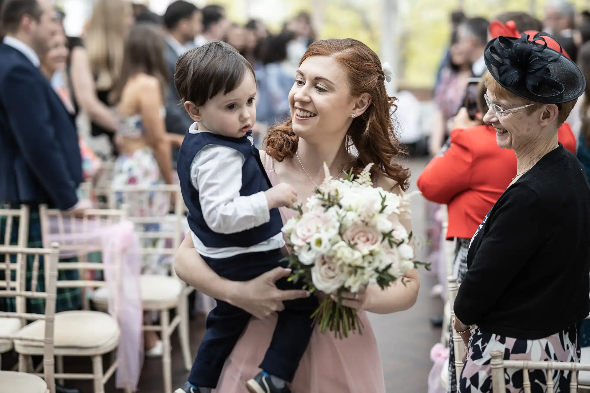 A woman in a pink dress holds a bouquet and a child. Another woman in a red jacket and hat smiles nearby. People are seated in the background.