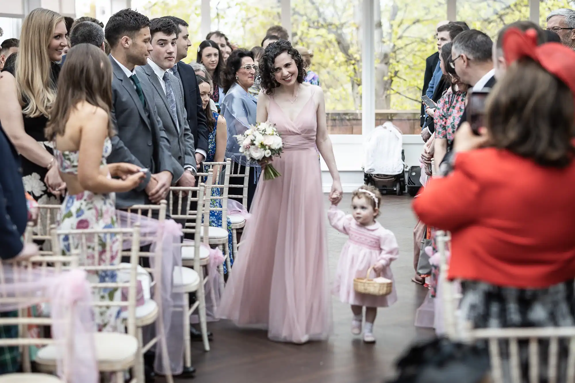 A woman in a pink dress walks down an aisle holding a bouquet, accompanied by a child in a similar dress carrying a basket. They are surrounded by seated and standing guests.