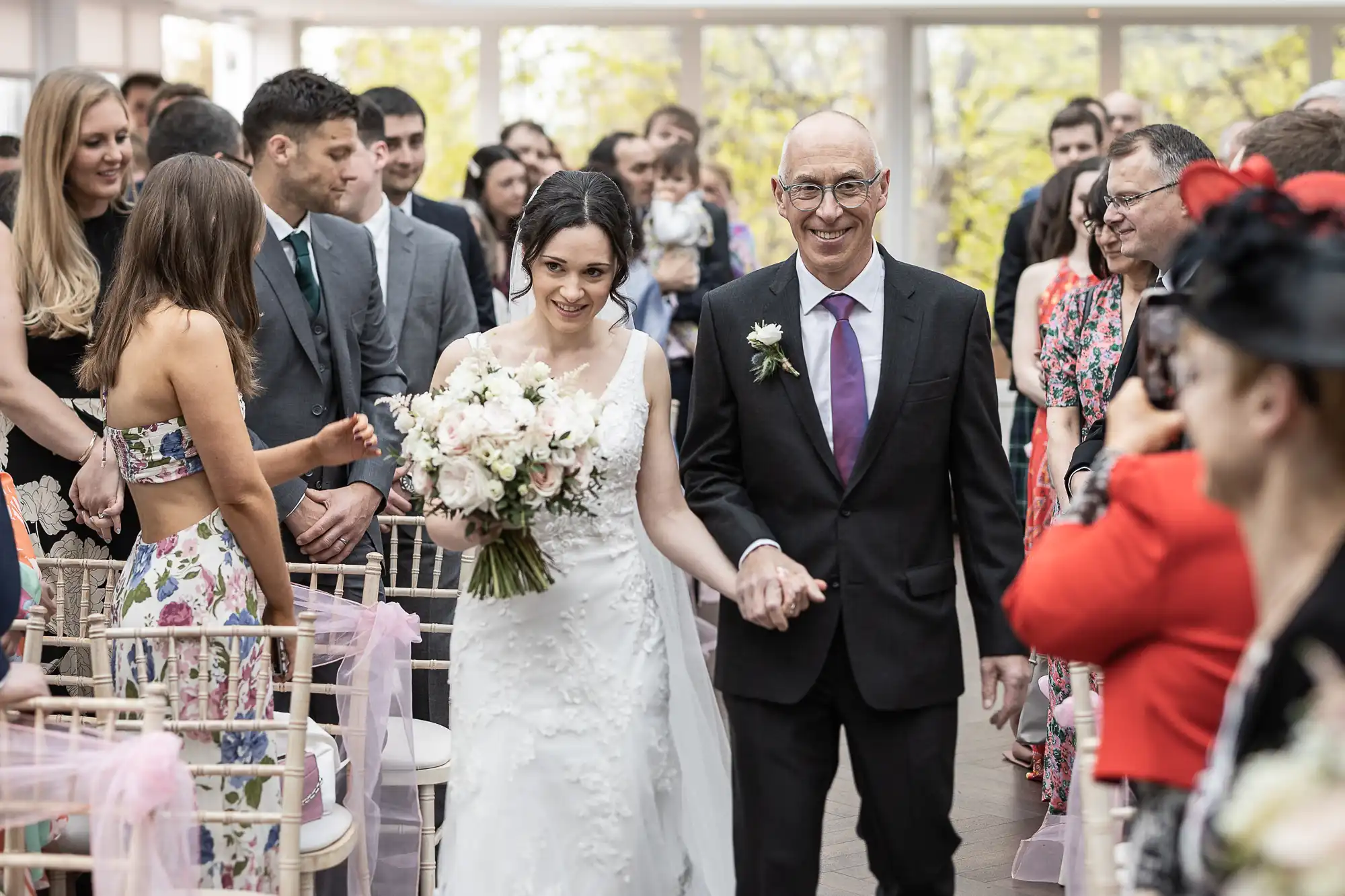 A bride in a white dress holds a bouquet and walks down the aisle with an older man in a suit, surrounded by seated guests in a bright room.