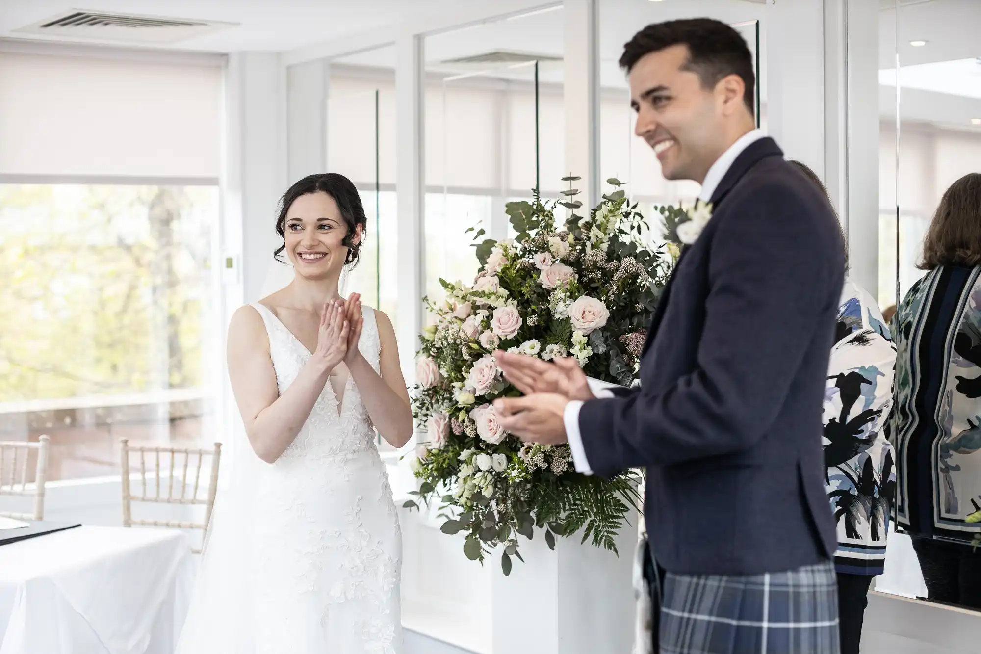 A bride and groom are clapping and smiling in a bright room, standing next to a large floral arrangement.