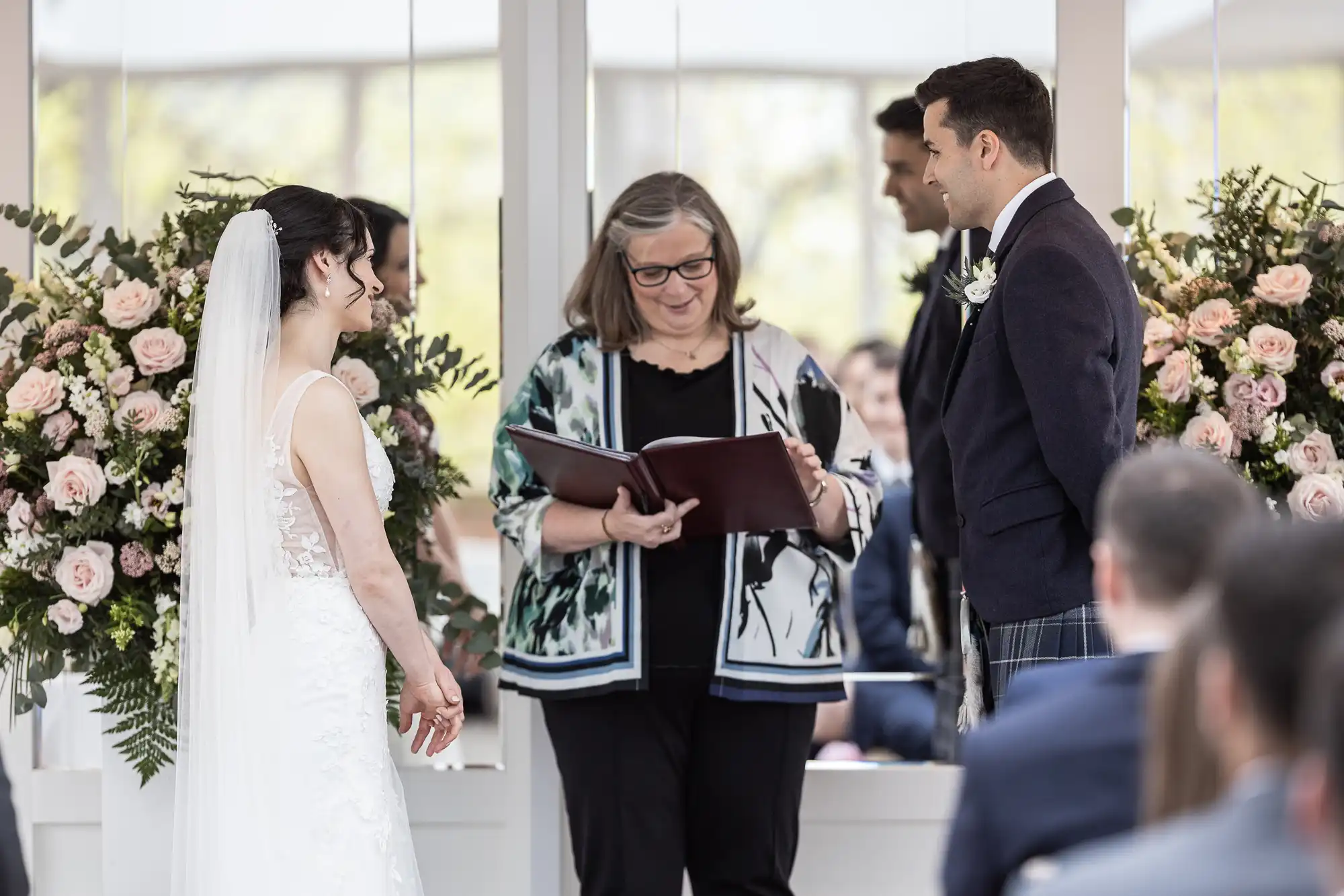 A bride and groom stand facing each other during a wedding ceremony indoors, with a woman officiating and holding a book. Floral arrangements are in the background.