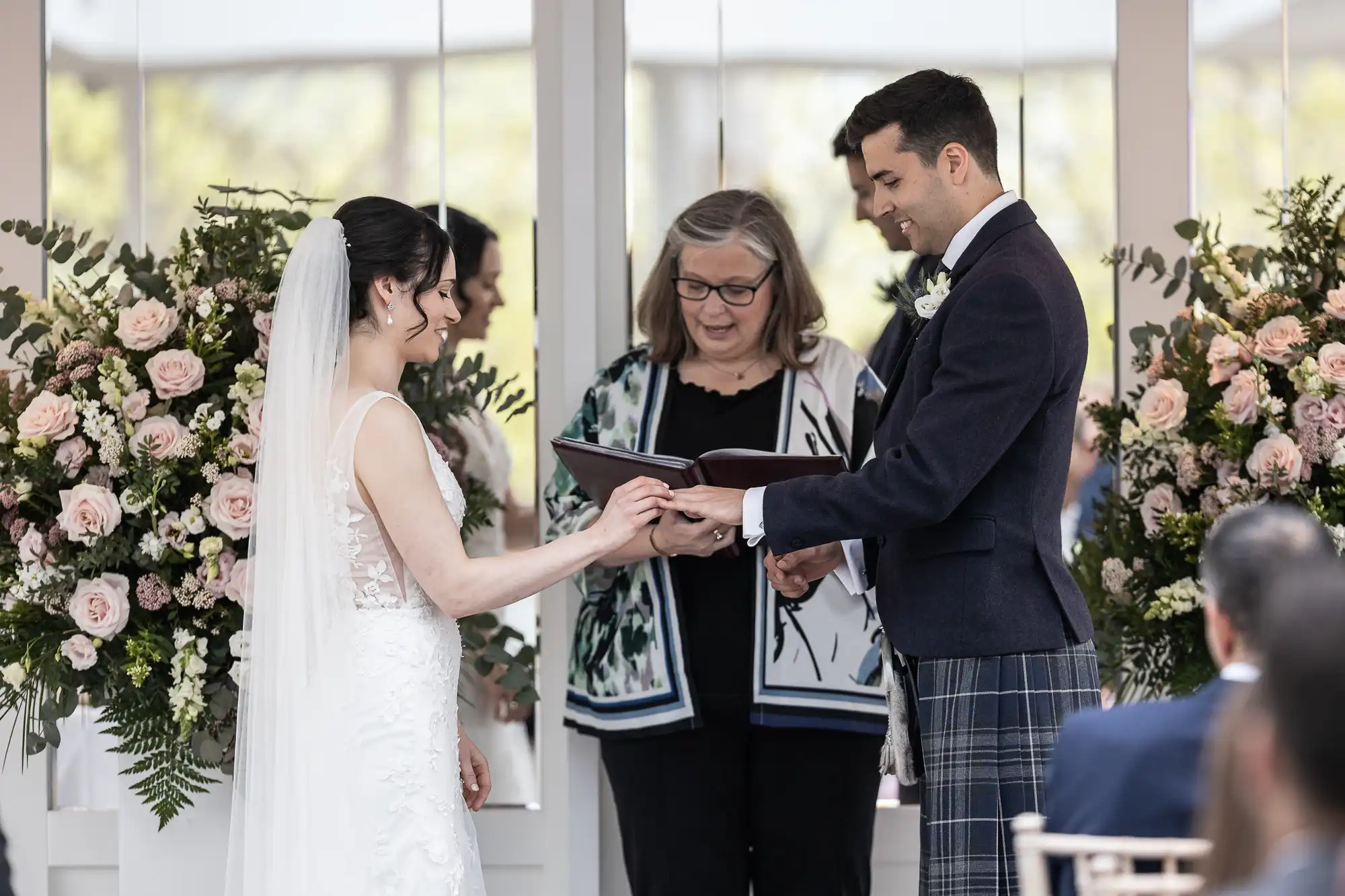 A couple exchanges vows during a wedding ceremony, with an officiant standing between them. They are surrounded by floral arrangements.
