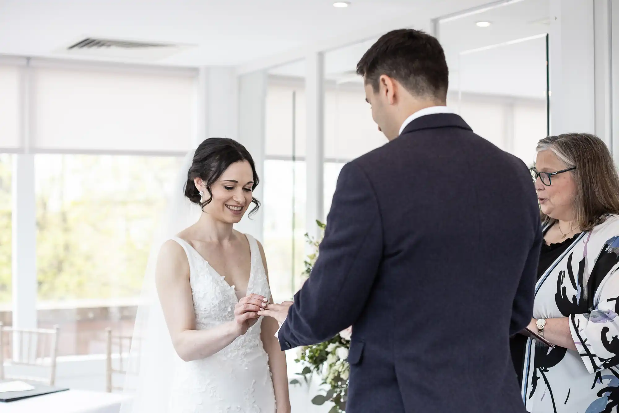 Bride and groom exchanging rings during a wedding ceremony, with an officiant standing nearby in a bright room.