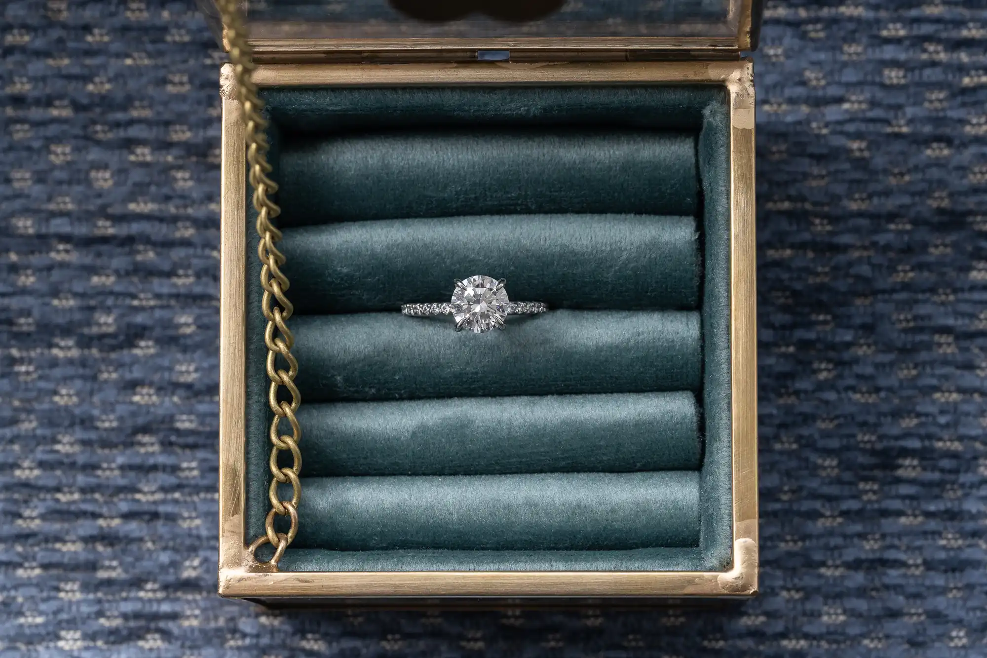 A diamond ring with a silver band is displayed in a textured dark green ring box on a blue and black patterned surface, with a gold chain attached to the box lid.