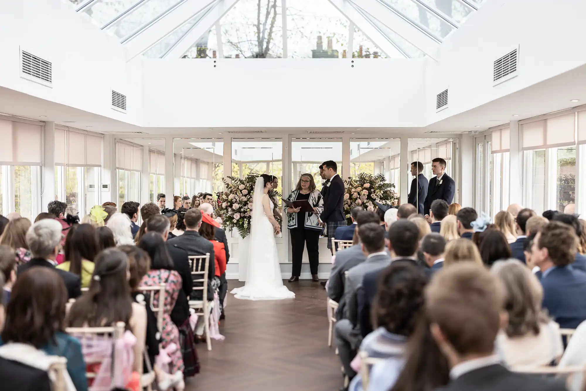 A bride and groom stand at an indoor wedding ceremony with a large gathering of seated guests. A person officiates in the center between them, surrounded by floral arrangements.