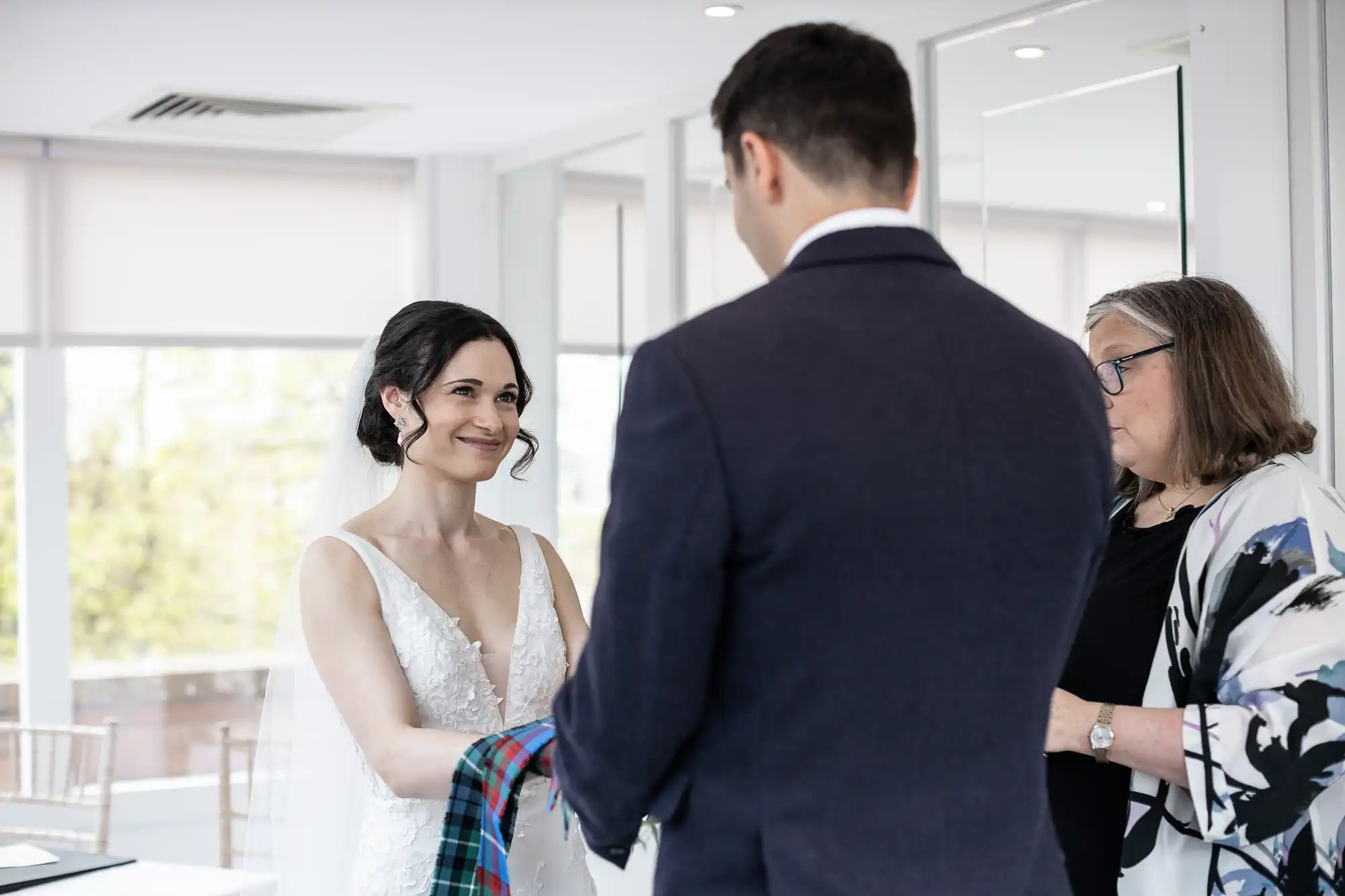 A bride and groom participate in a handfasting ceremony, holding a tartan cloth. A woman stands nearby reading. The setting appears to be a brightly lit room with large windows.