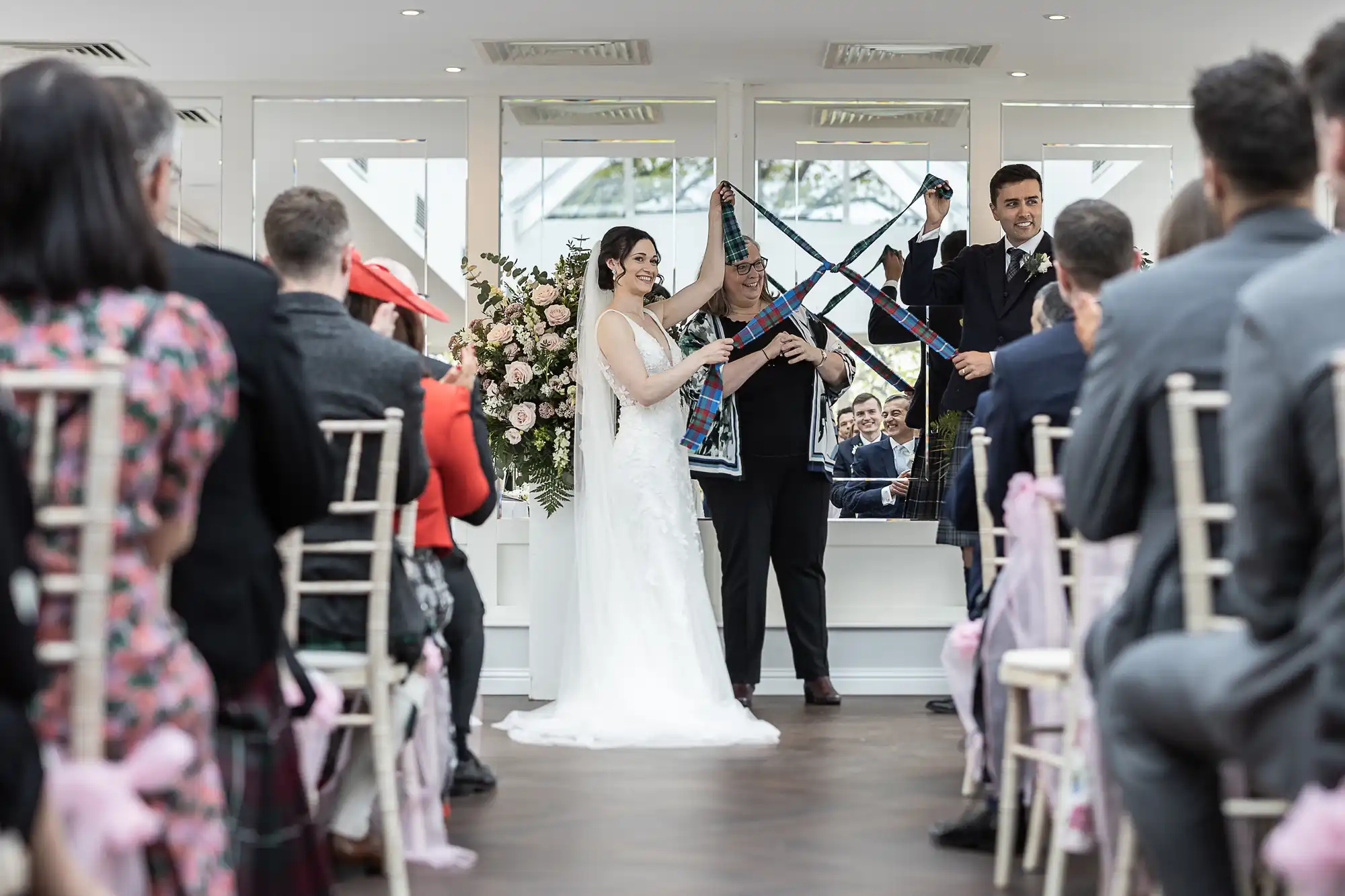 Bride and groom participate in a symbolic wedding ritual with ribbon tying, surrounded by guests in a bright ceremony space.