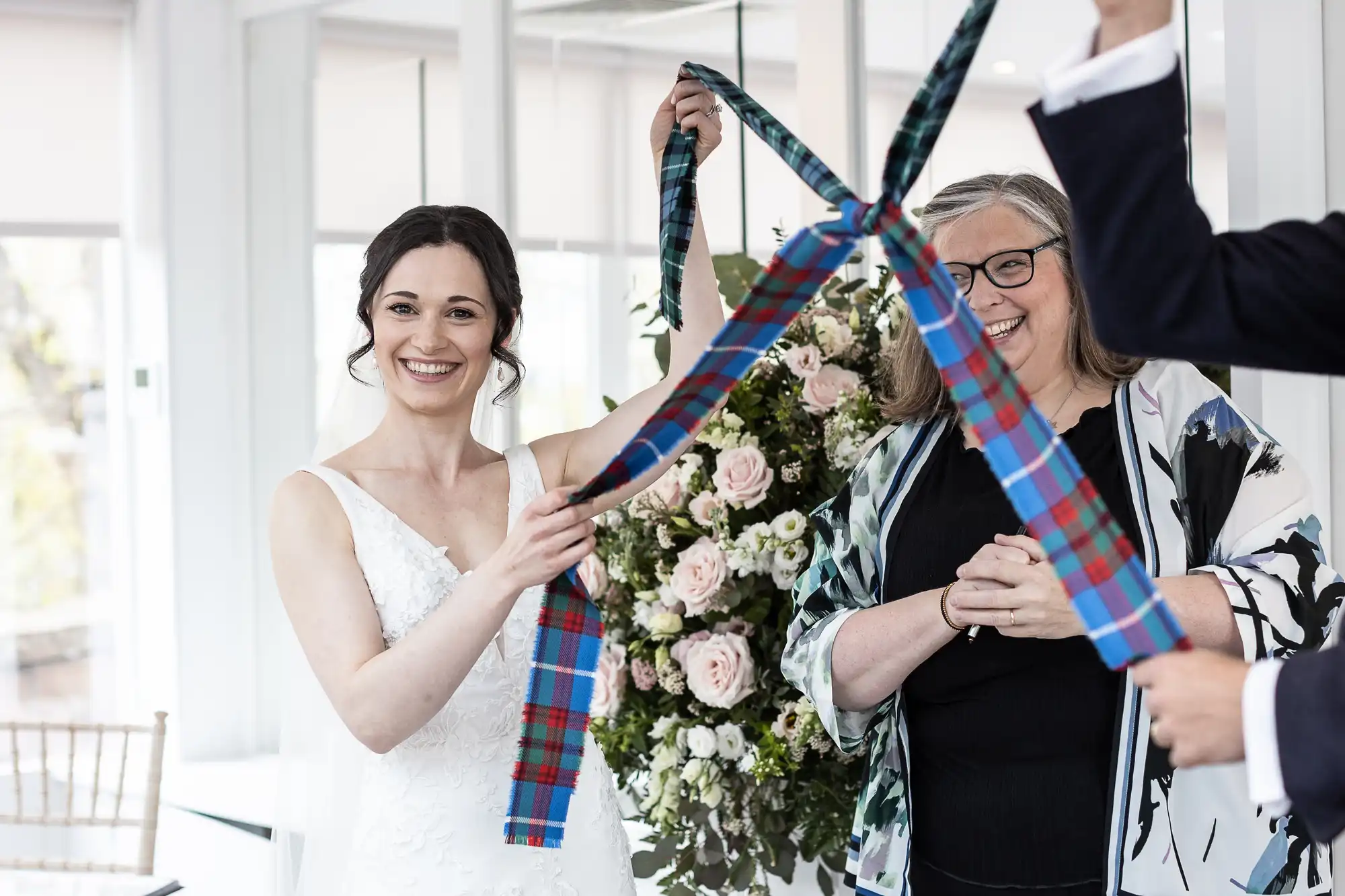 Bride smiling while holding a plaid ribbon tied to a flower arrangement, with a woman standing beside her. Other hands hold the ribbon.