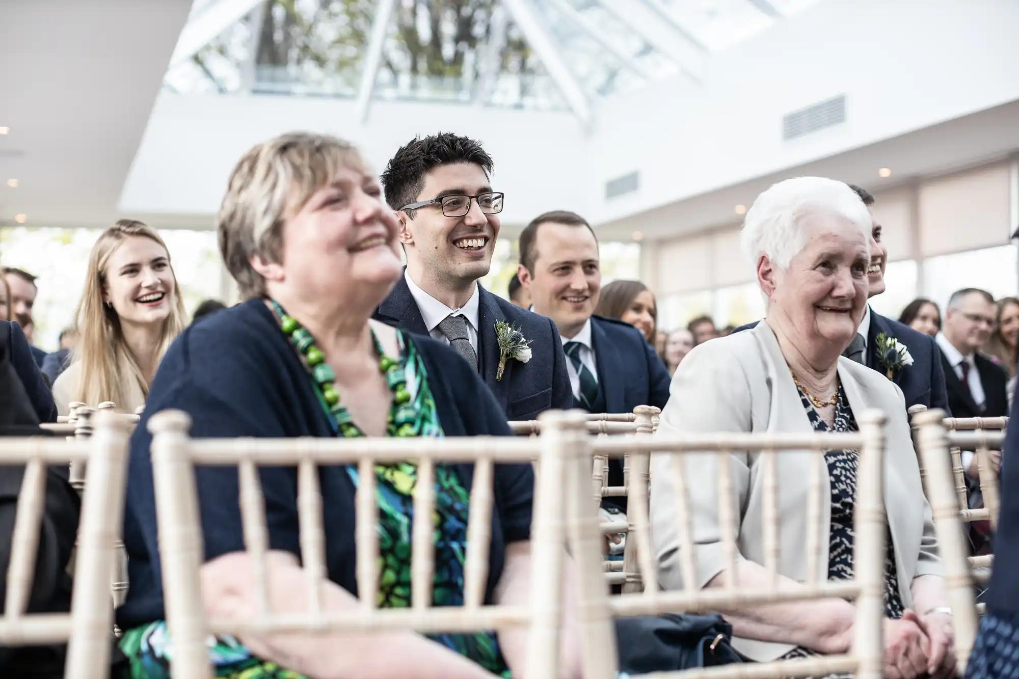 An audience of people, including seniors and younger adults, are sitting on wooden chairs and smiling at an indoor event.