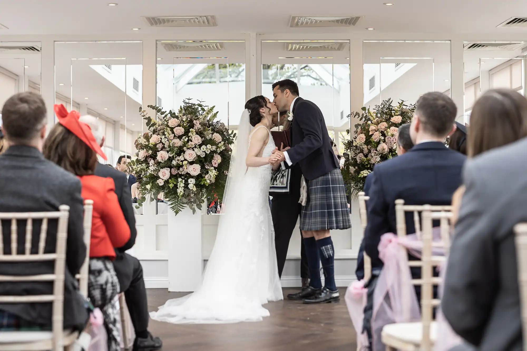 Bride and groom kiss at their wedding ceremony, surrounded by guests seated on either side, with large floral arrangements in the background.