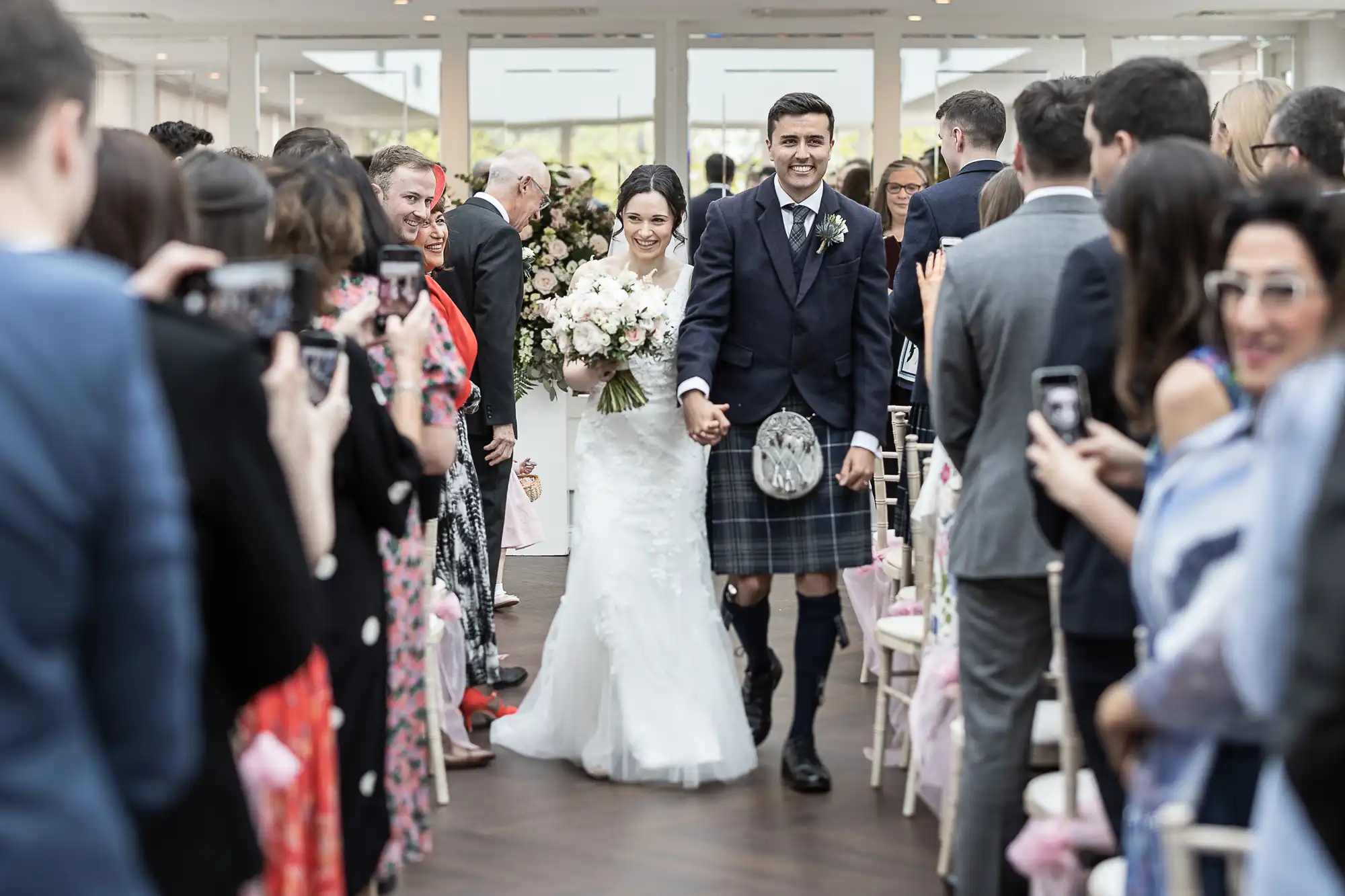 Bride in white gown and groom in kilt walking down the aisle, surrounded by guests taking photos.