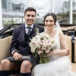 A bride and groom sit together in a vehicle, smiling at the camera. The bride holds a bouquet of flowers, and the groom is wearing a kilt.