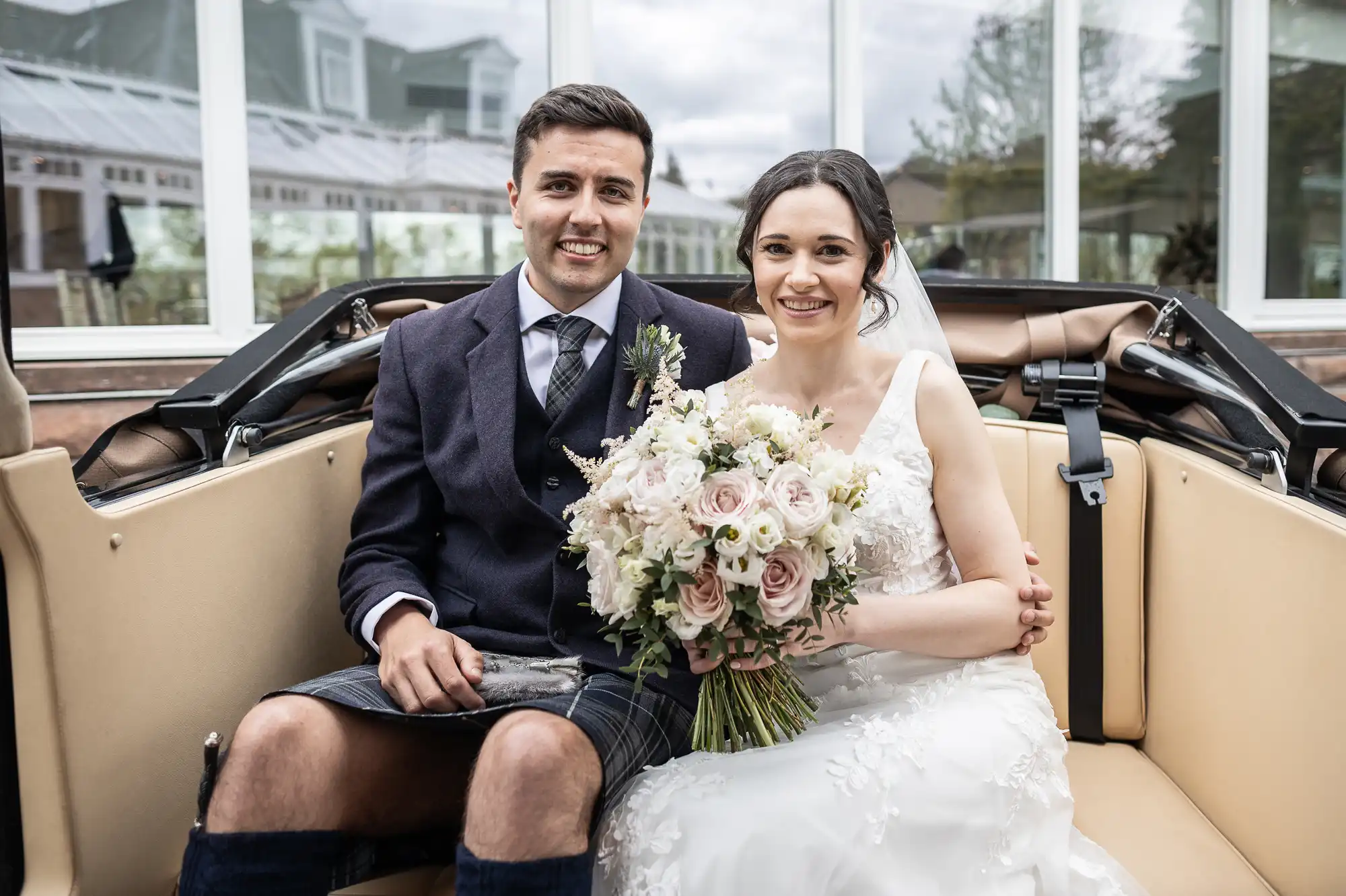 A bride and groom sit together in a vehicle, smiling at the camera. The bride holds a bouquet of flowers, and the groom is wearing a kilt.