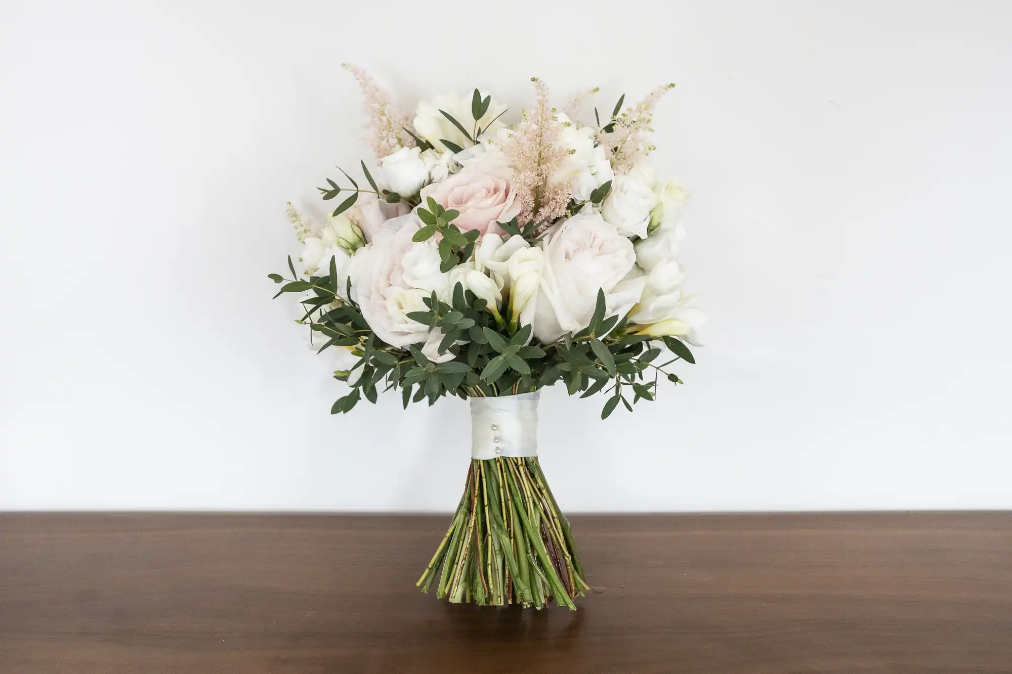 A bouquet of white and pale pink flowers with greenery, wrapped in white ribbon, placed on a wooden surface against a plain white background.